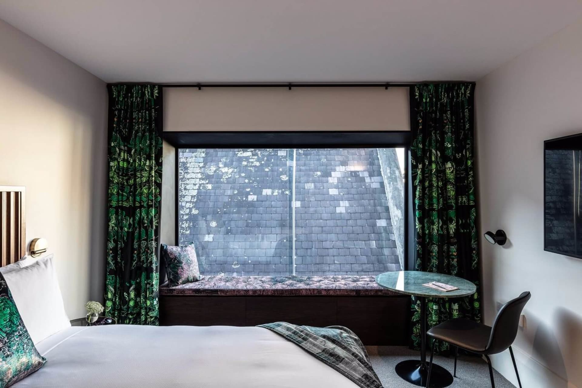 A modern minimalist bedroom featuring a bed with a wooden headboard and white linens, a small table with a black top and chai