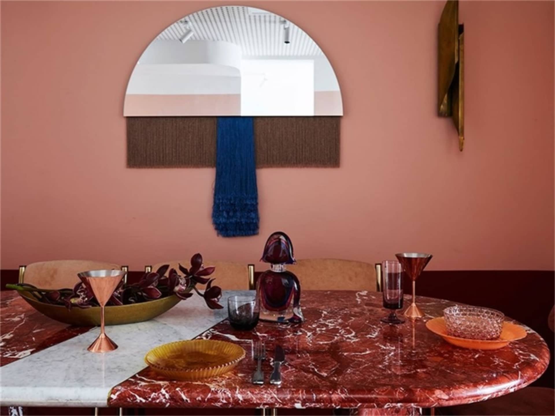 A modern dining room with a round, red marble table, and a wall mirror with a blue tassel decoration.