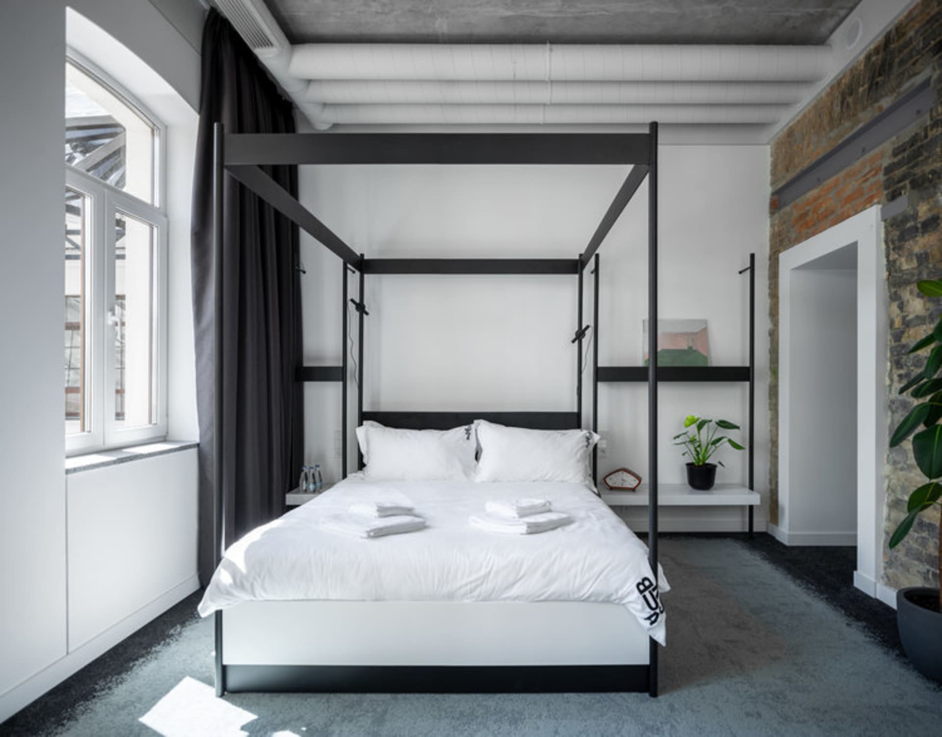 極簡臥室內設有混凝土與金屬材質的四柱床,搭配白色寢具與大型窗戶。
