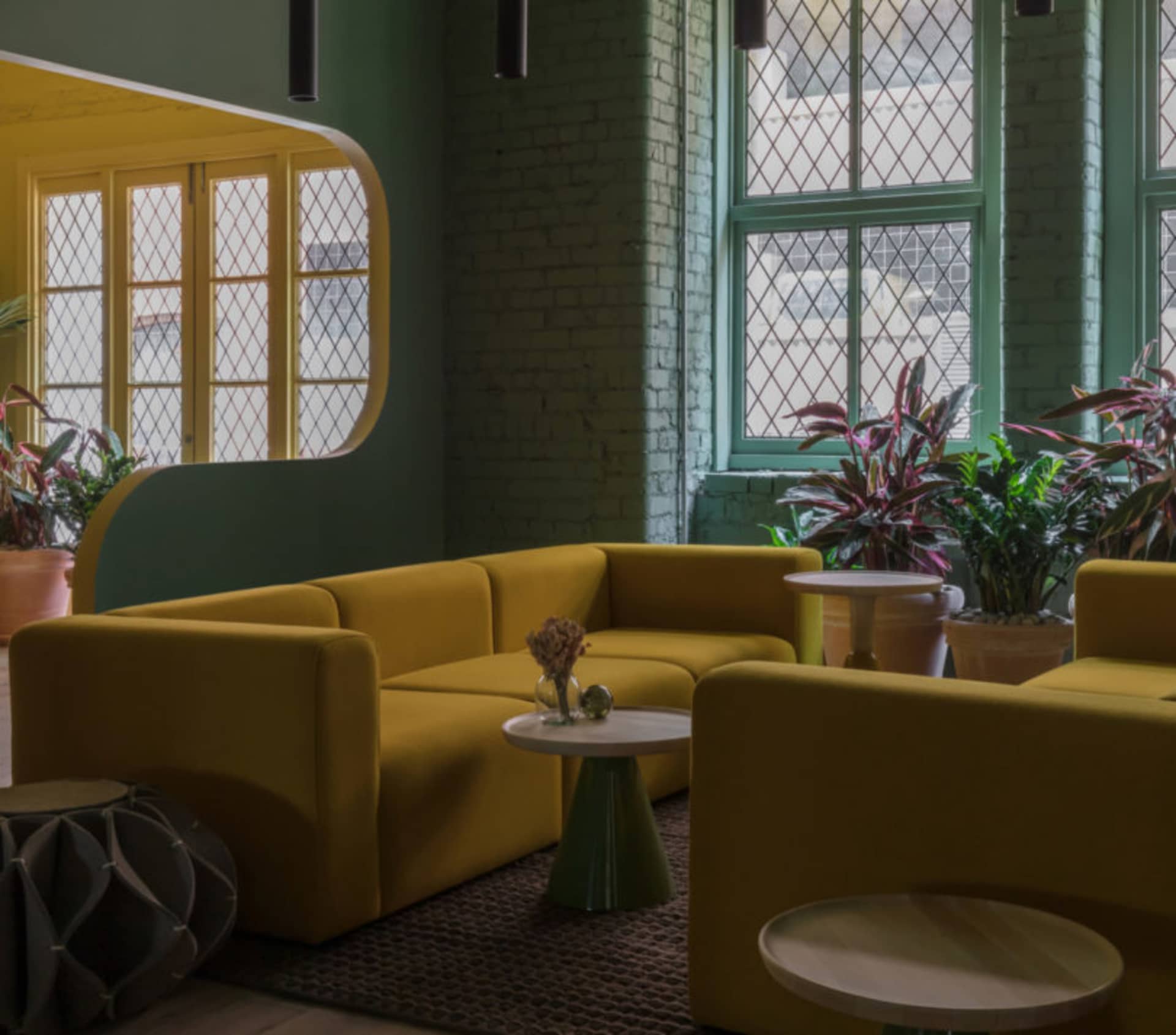 A modern living room with mustard-yellow upholstered furniture, green brick walls, and potted plants.
