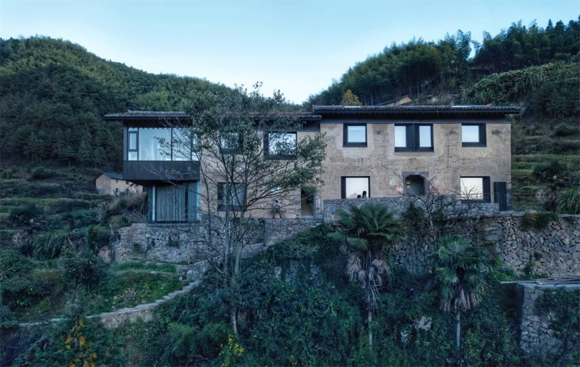 一座現代住宅建築，呈現一座梯田式石屋坐落於鬱鬱蔥蔥的綠色山坡上。