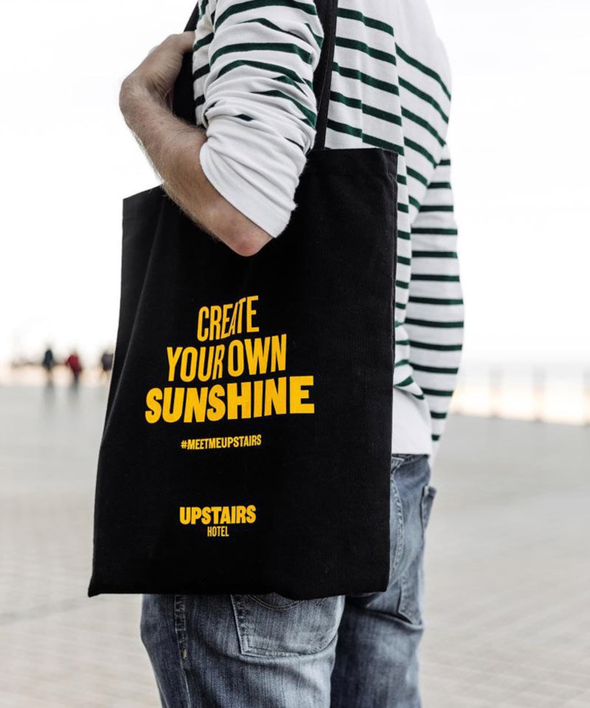 Person holding a black tote bag with 'CREATE YOUR OWN SUNSHINE' text on it.