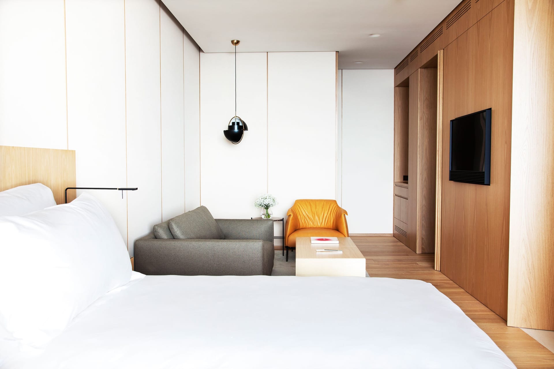 A minimalist bedroom featuring a white bed with a wooden headboard, a sofa, and an armchair, all against a background of whit