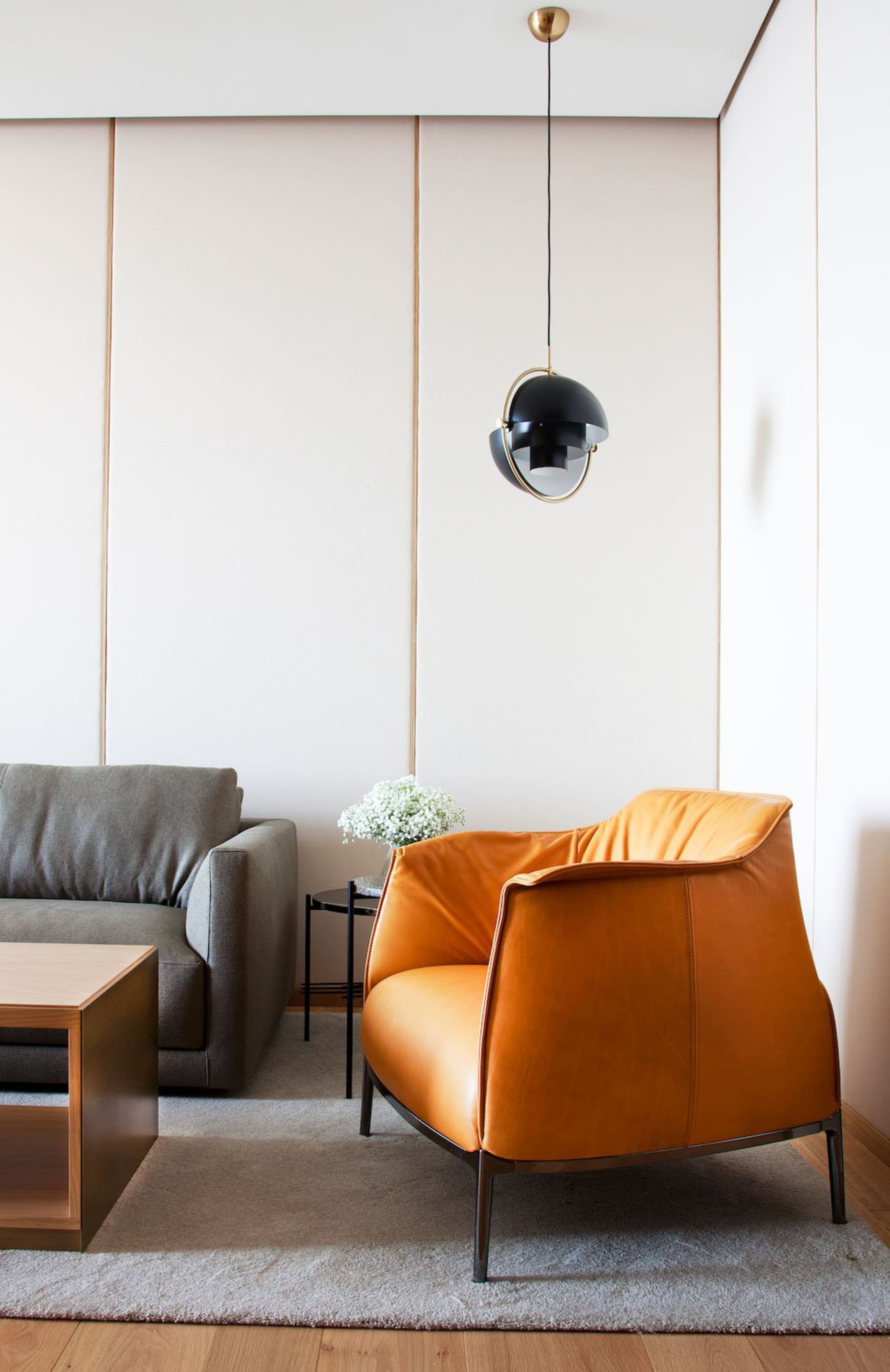 A modern living room features an orange leather armchair, a gray sofa, and a wooden coffee table in front of a white panel wa