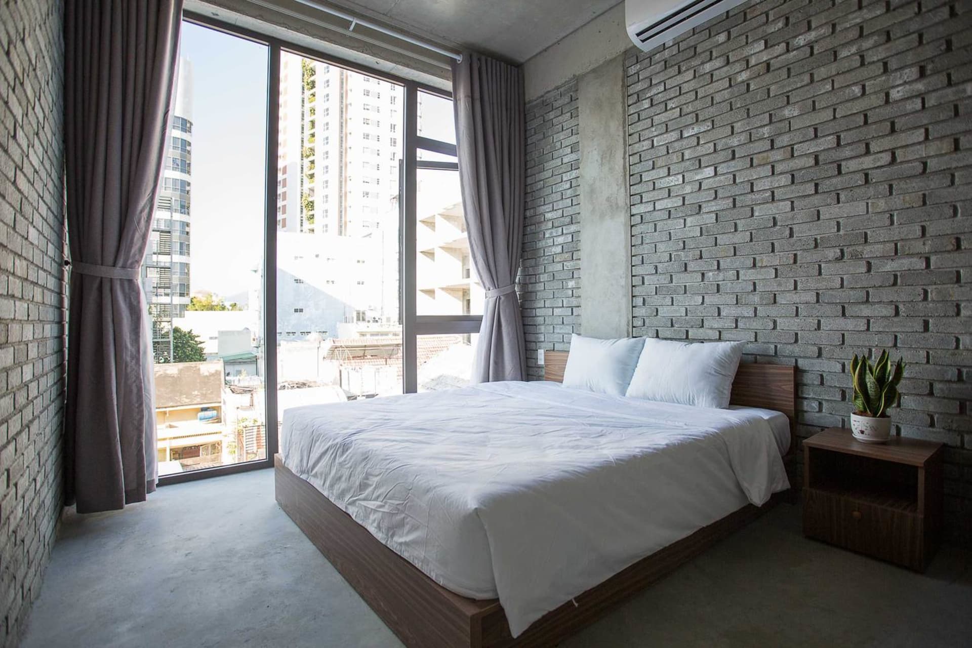 Modern minimalist bedroom with a bed, white bedding, grey curtains, and a brick wall