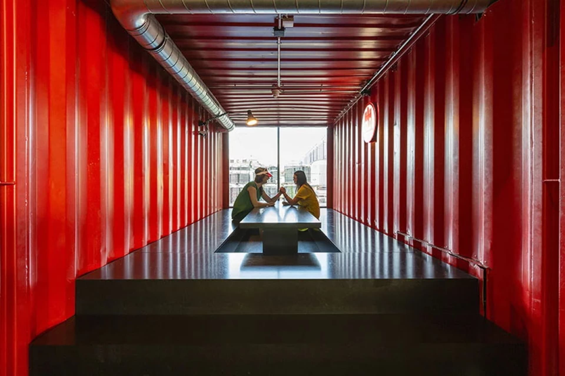 Two individuals seated at a table in a shipping container setting, surrounded by red metal walls.