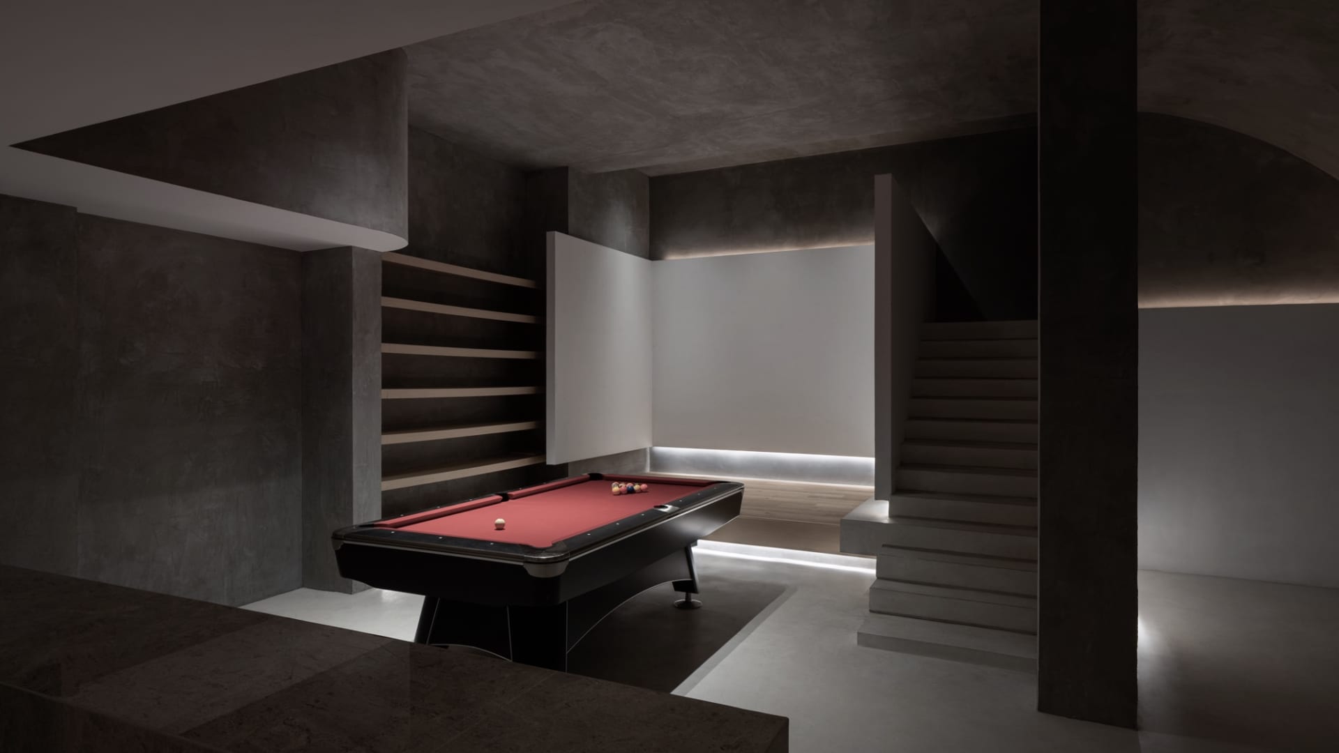 A minimalist, modern basement with a concrete floor, a red pool table, and a white wall with built-in shelves.