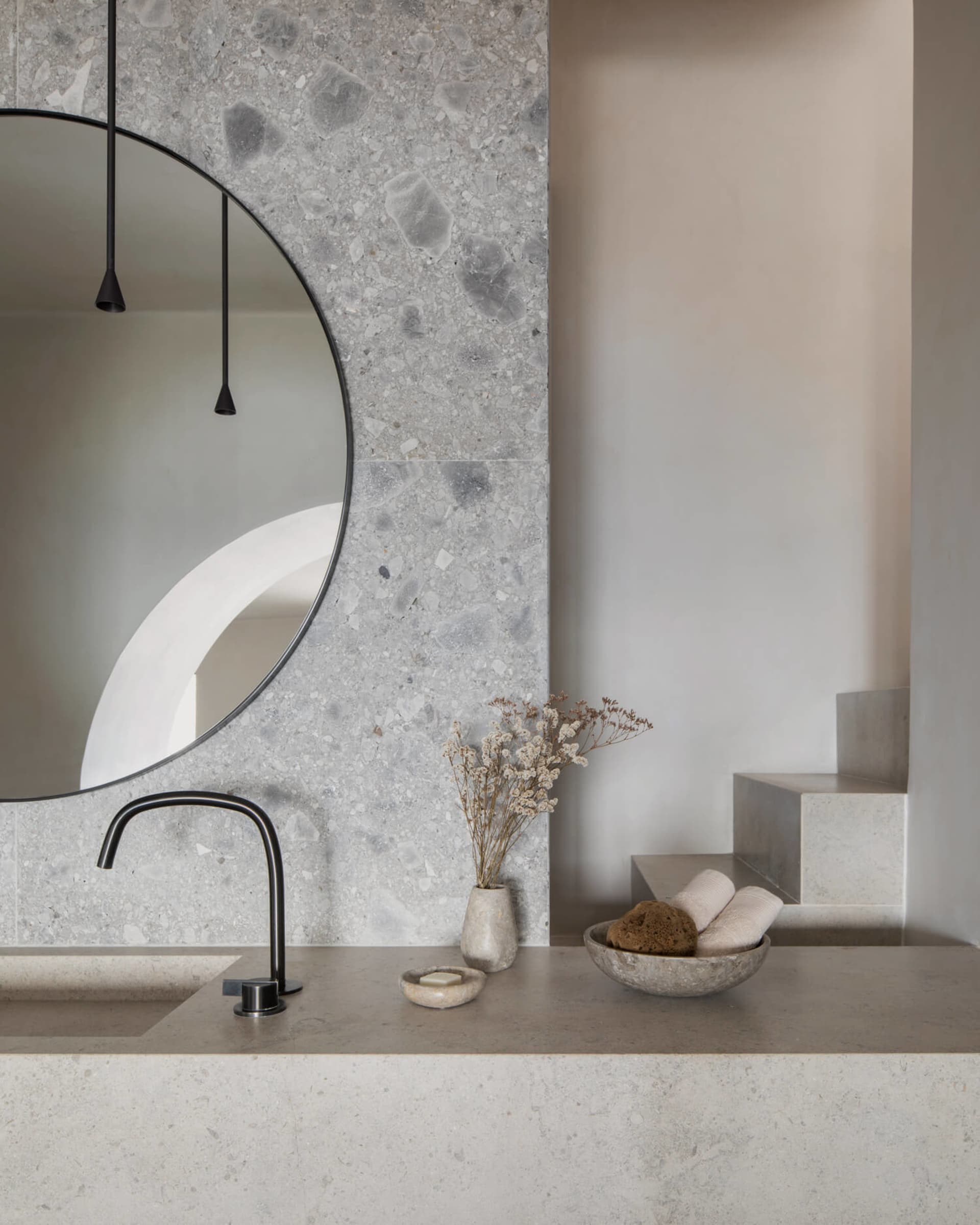 Modern minimalist bathroom featuring a circular mirror mounted on a terrazzo wall, a floating vanity, and dried flowers in a