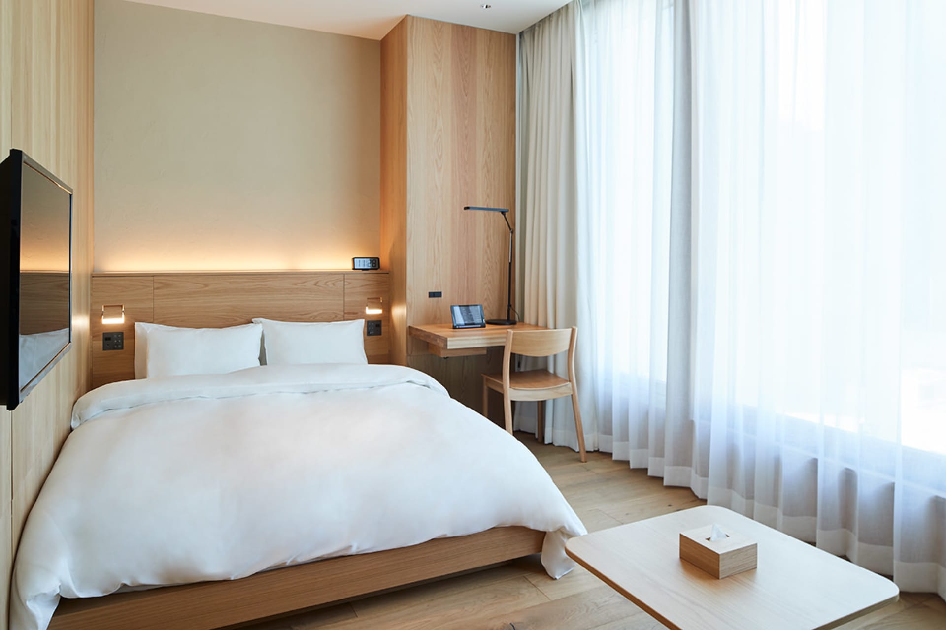 Modern minimalist hotel room with a wooden bed and glass window, featuring white textiles and a functional desk area.