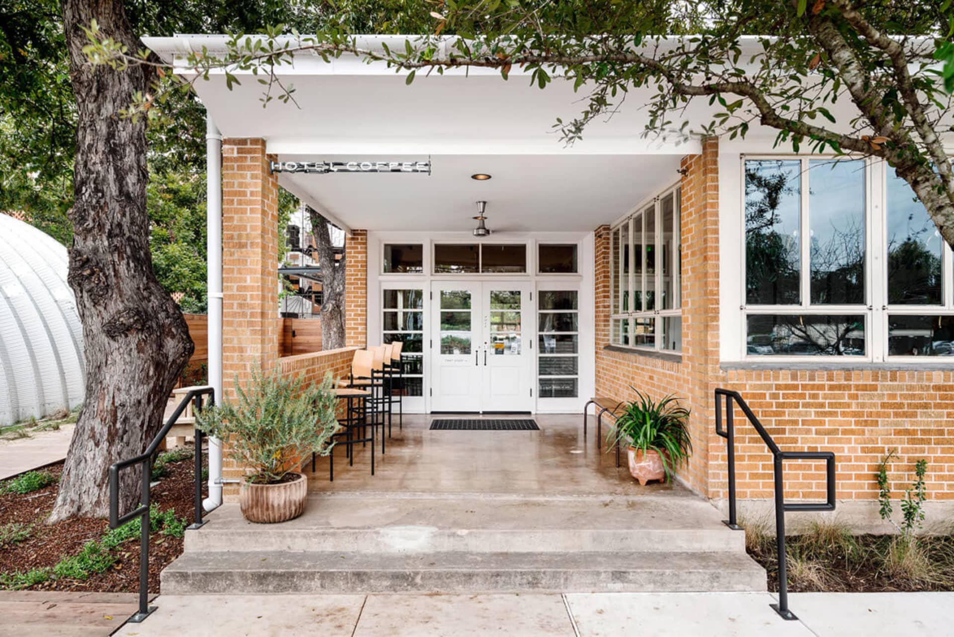 The exterior of a cafe with white brick walls, concrete flooring, and potted plants on steps.