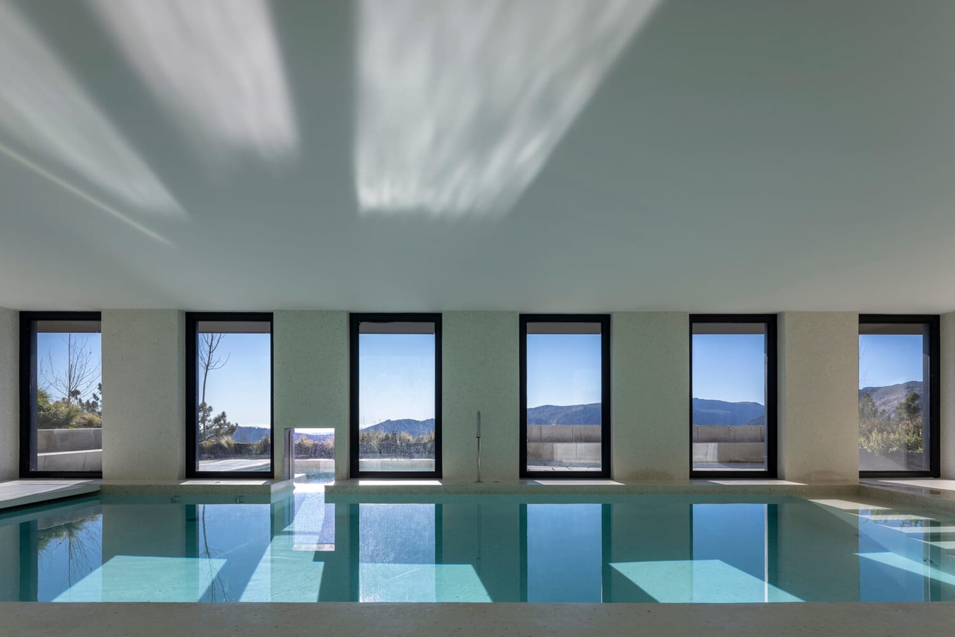 Modern industrial-style indoor swimming pool with concrete walls and floor, featuring large windows overlooking scenic mounta