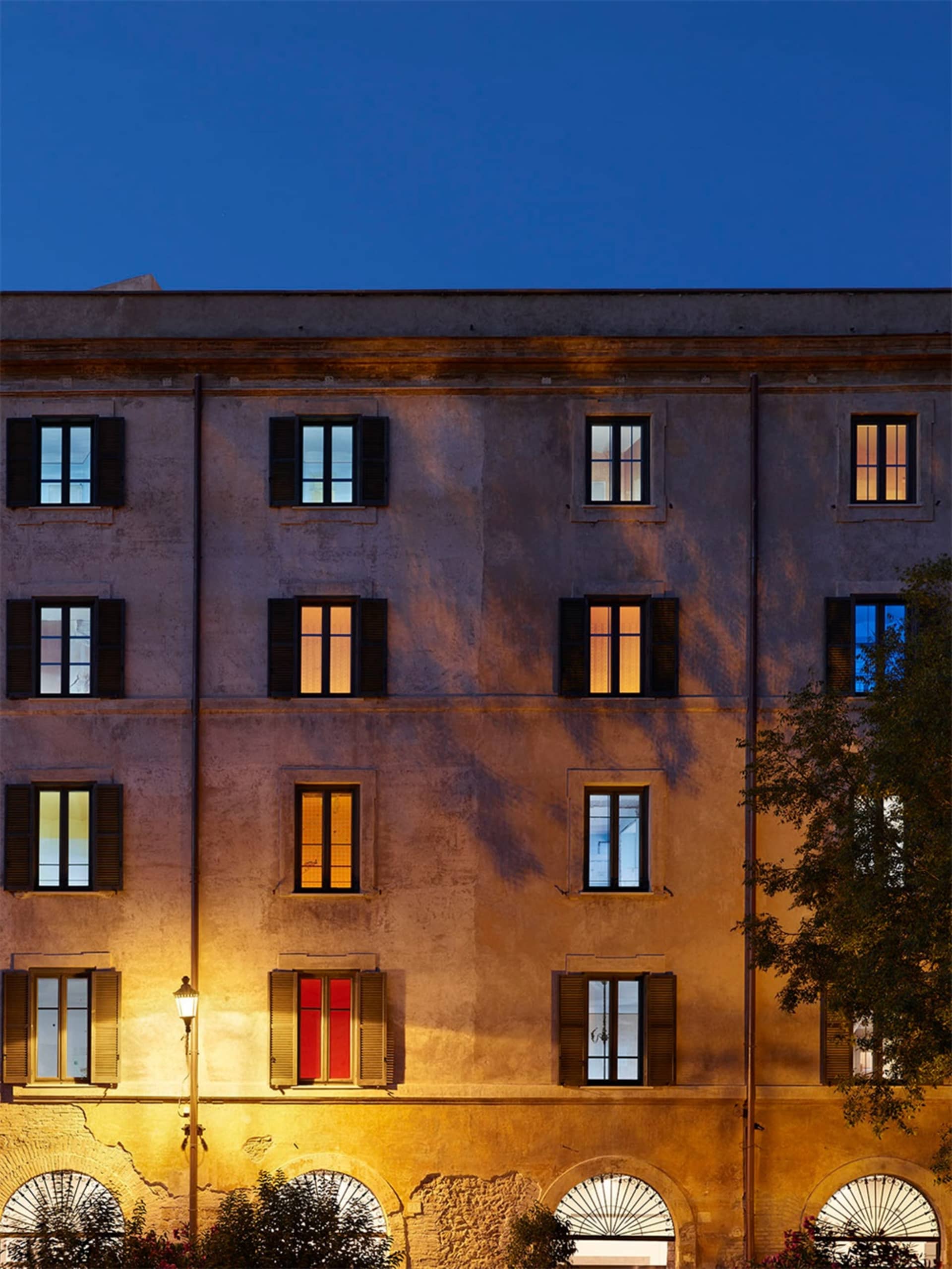 A stone building facade illuminated at dusk with lights on inside several windows.