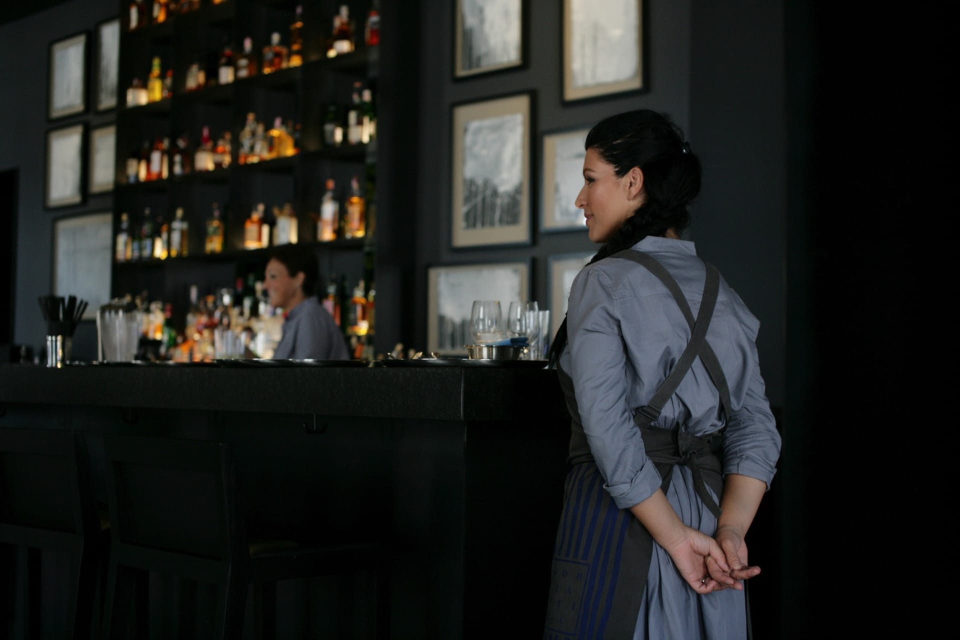A woman wearing an apron standing at a bar with a backdrop of shelves filled with bottles and framed pictures on the wall.