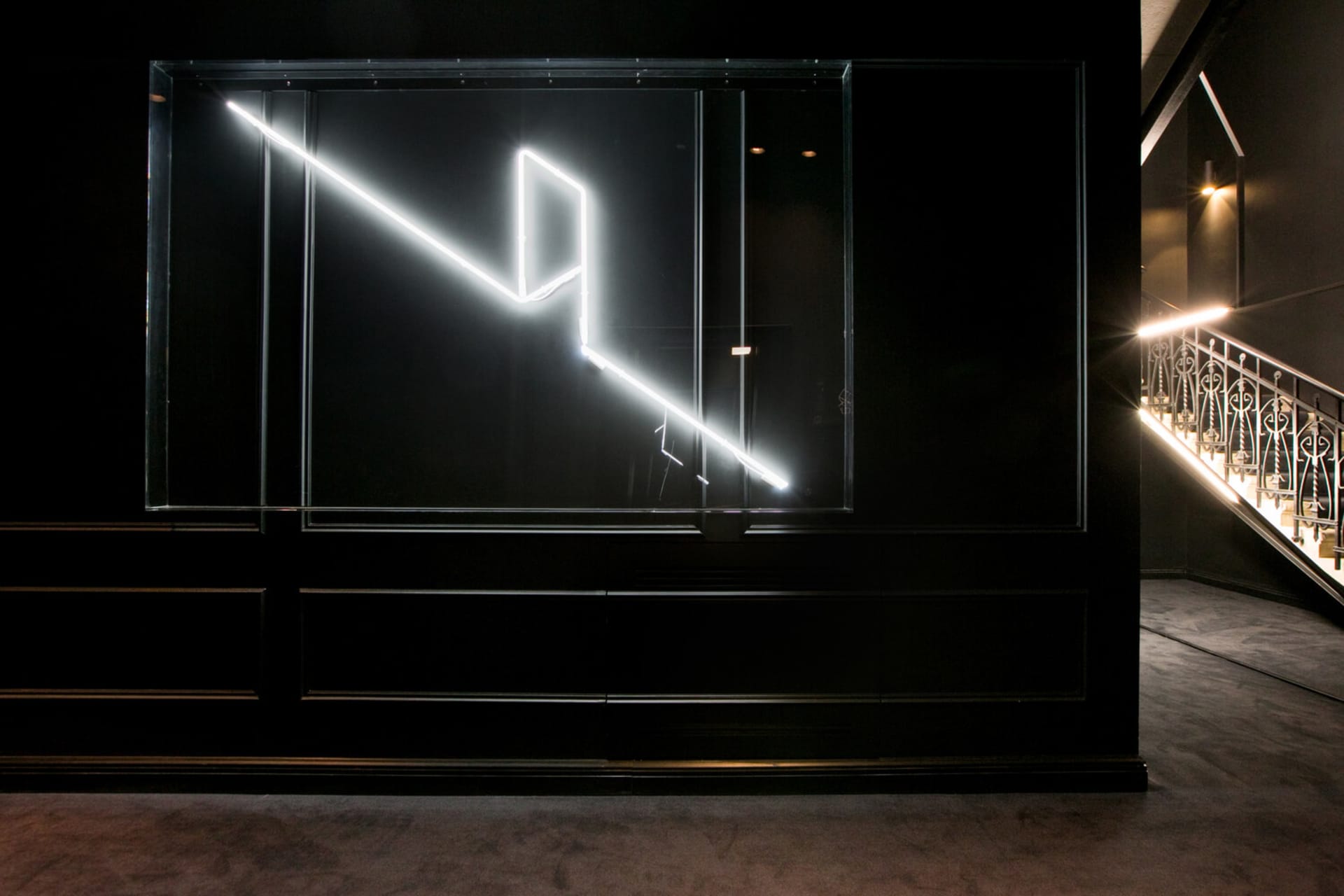 A minimalist installation art piece featuring a neon light sculpture mounted on a black wall.