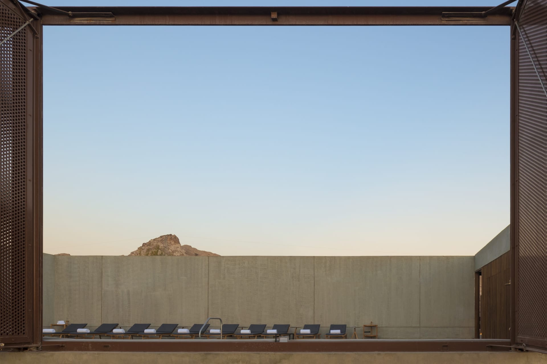 A minimalist poolside area with concrete walls, featuring several lounge chairs lined up under a clear blue sky.