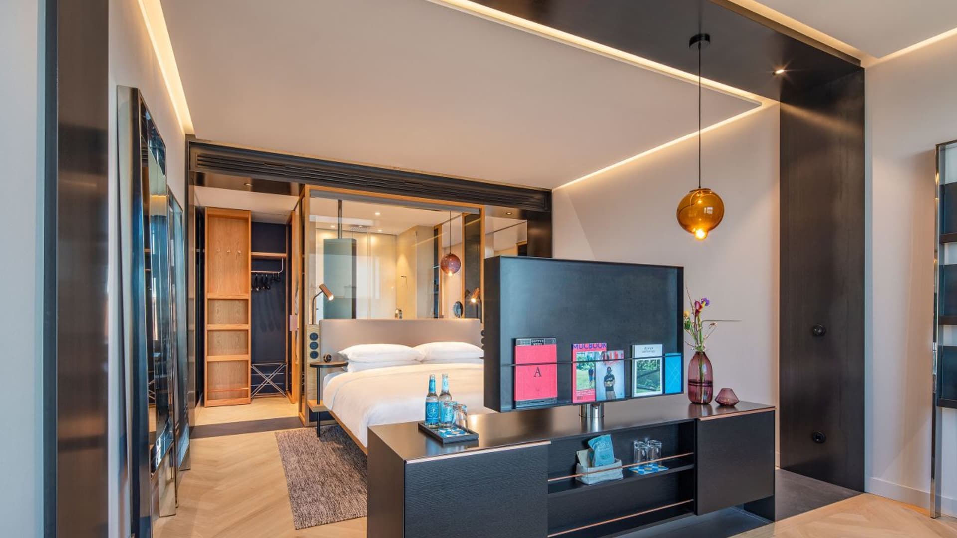 Modern hotel room with wooden flooring and glass walls, featuring a bed, nightstand, and magazines.