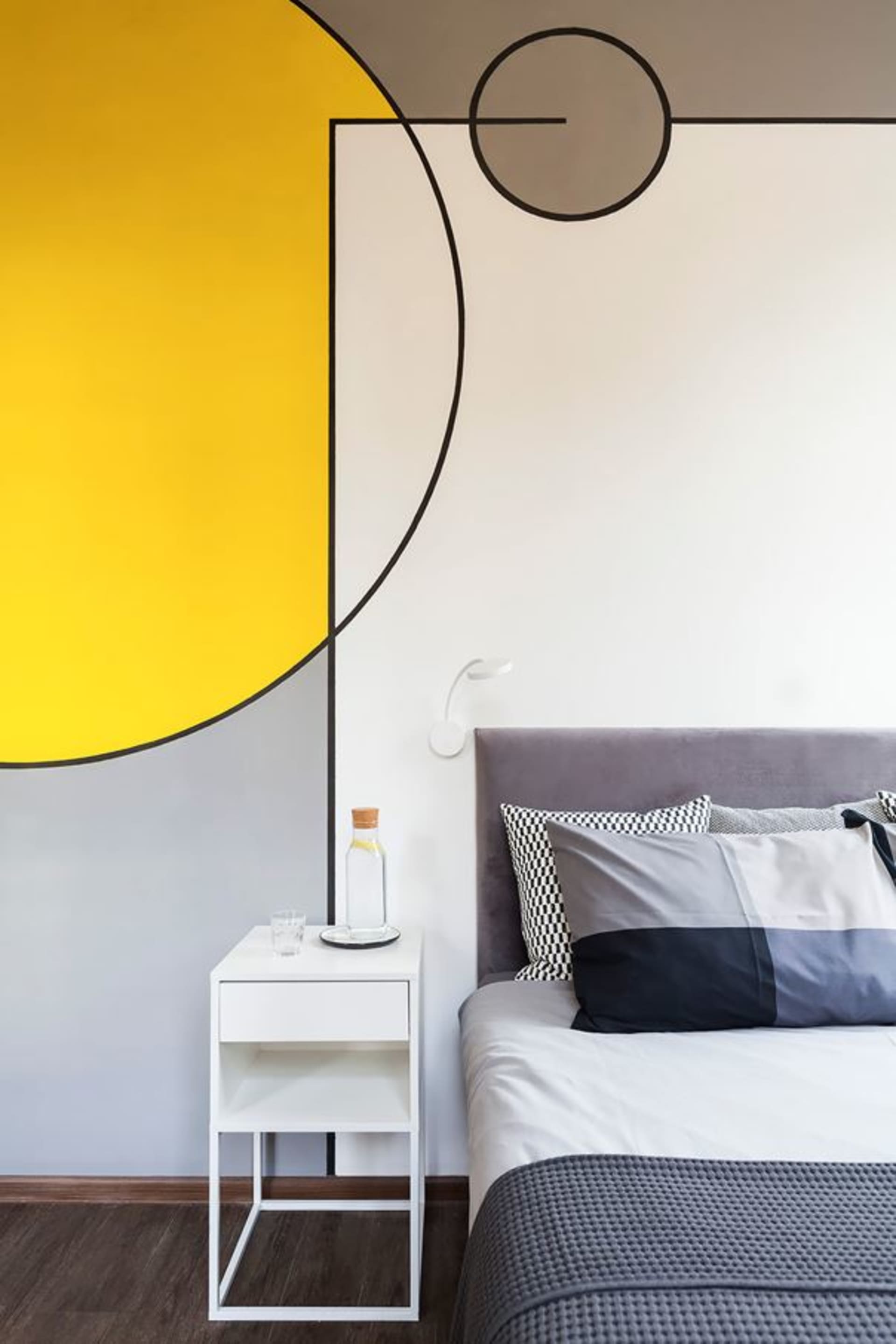A modern minimalist bedroom featuring a grey upholstered bed, a white metal nightstand with a small glass and a cork bottle,
