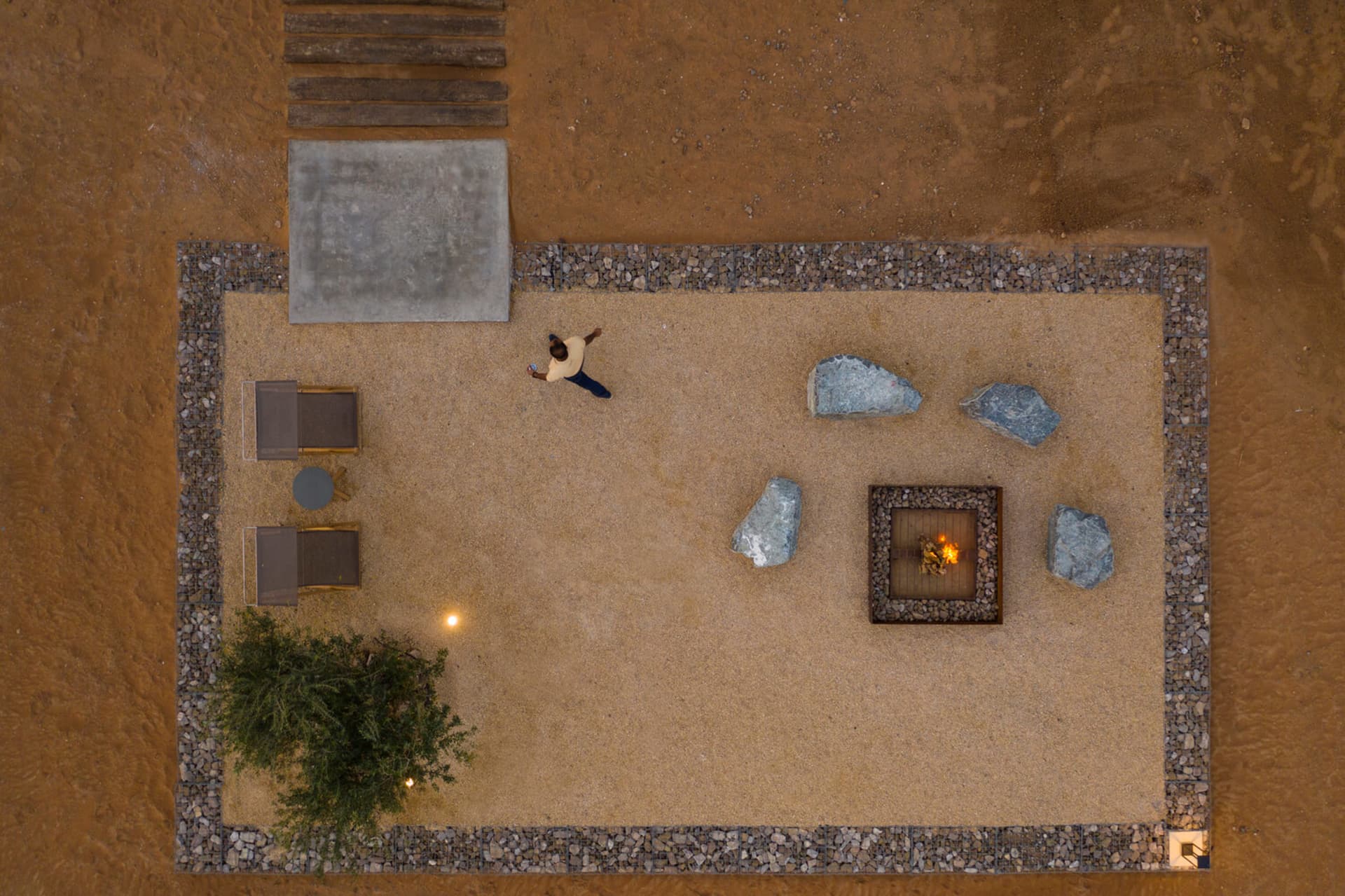 A desert outdoor living space with a fire pit, rocks, gravel, and a person walking nearby.