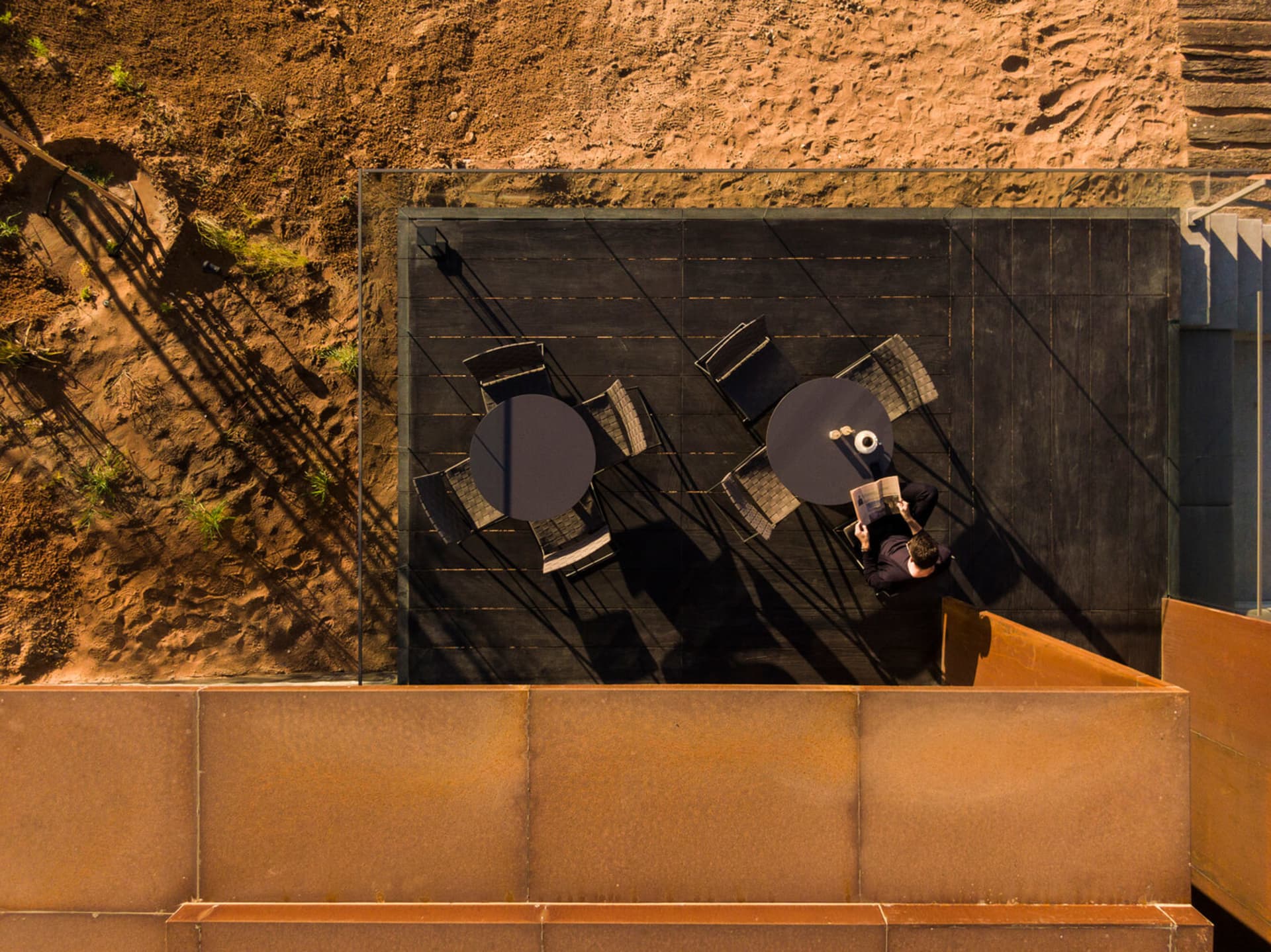 A modern terrace scene featuring sandstone, concrete, and wood materials, with a person reading a book at a table.