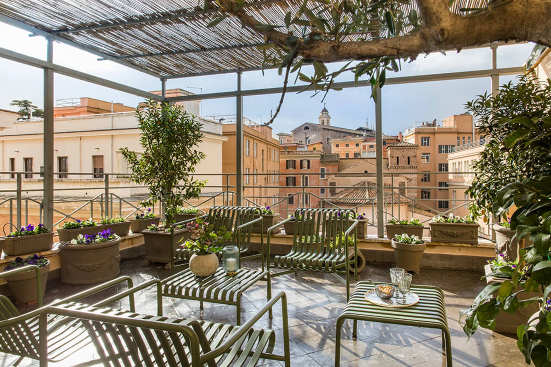 A rooftop patio with metal chairs and tables surrounded by potted plants, set against a backdrop of buildings and a church sp