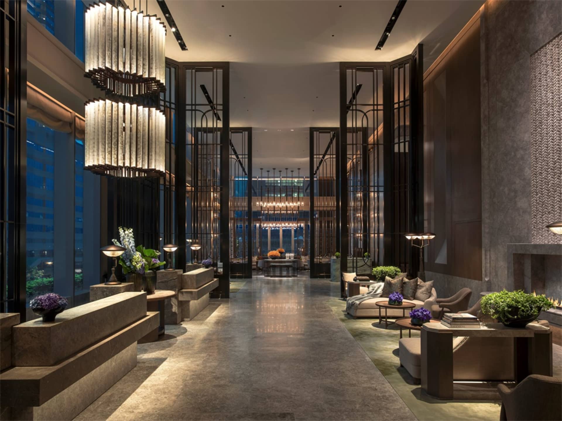 Modern lobby featuring metal light fixtures, wooden benches, glass panels, and stone flooring.