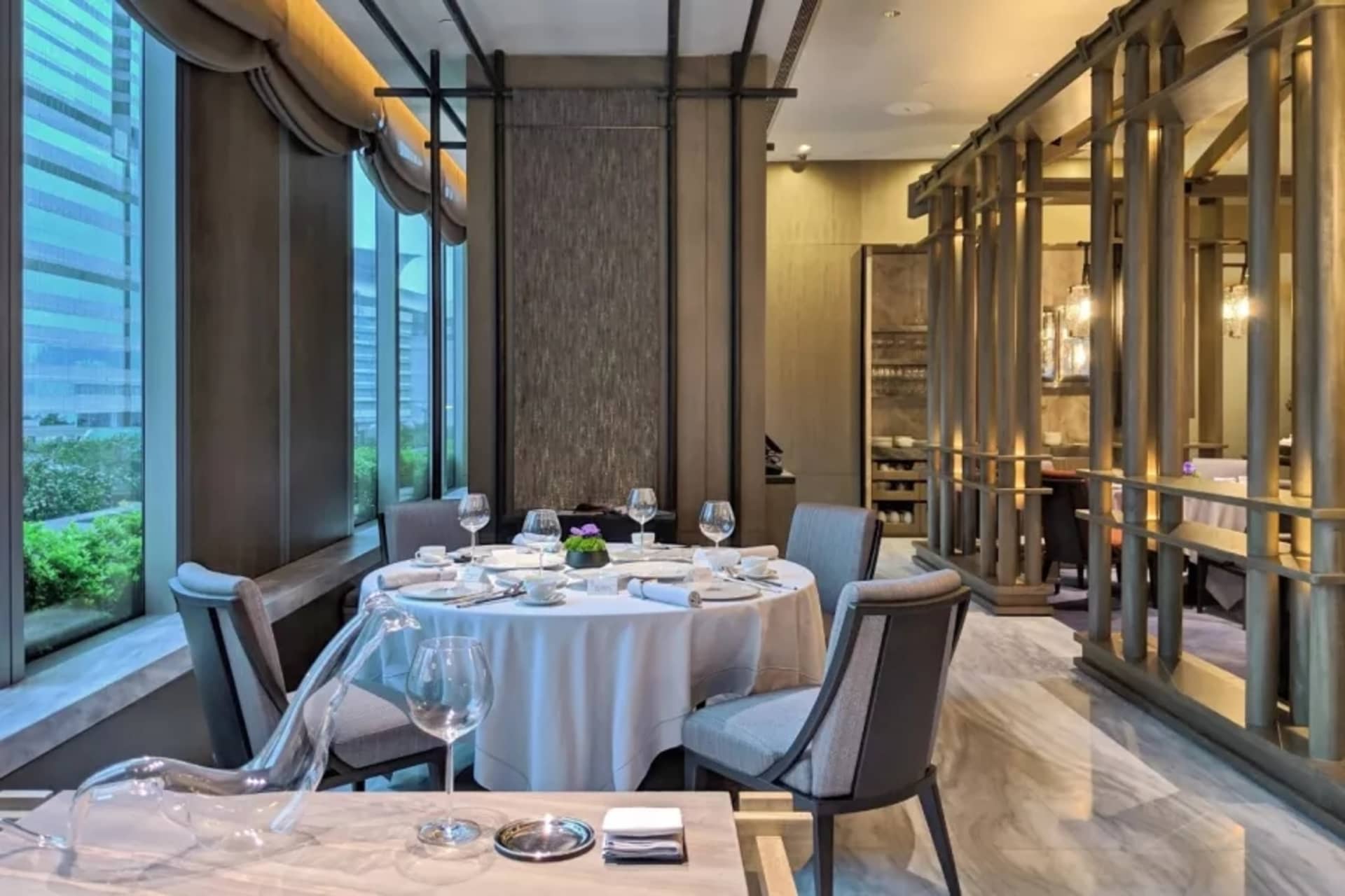 Modern restaurant dining room with marble flooring, metal railings, and floor-to-ceiling windows overlooking cityscape.