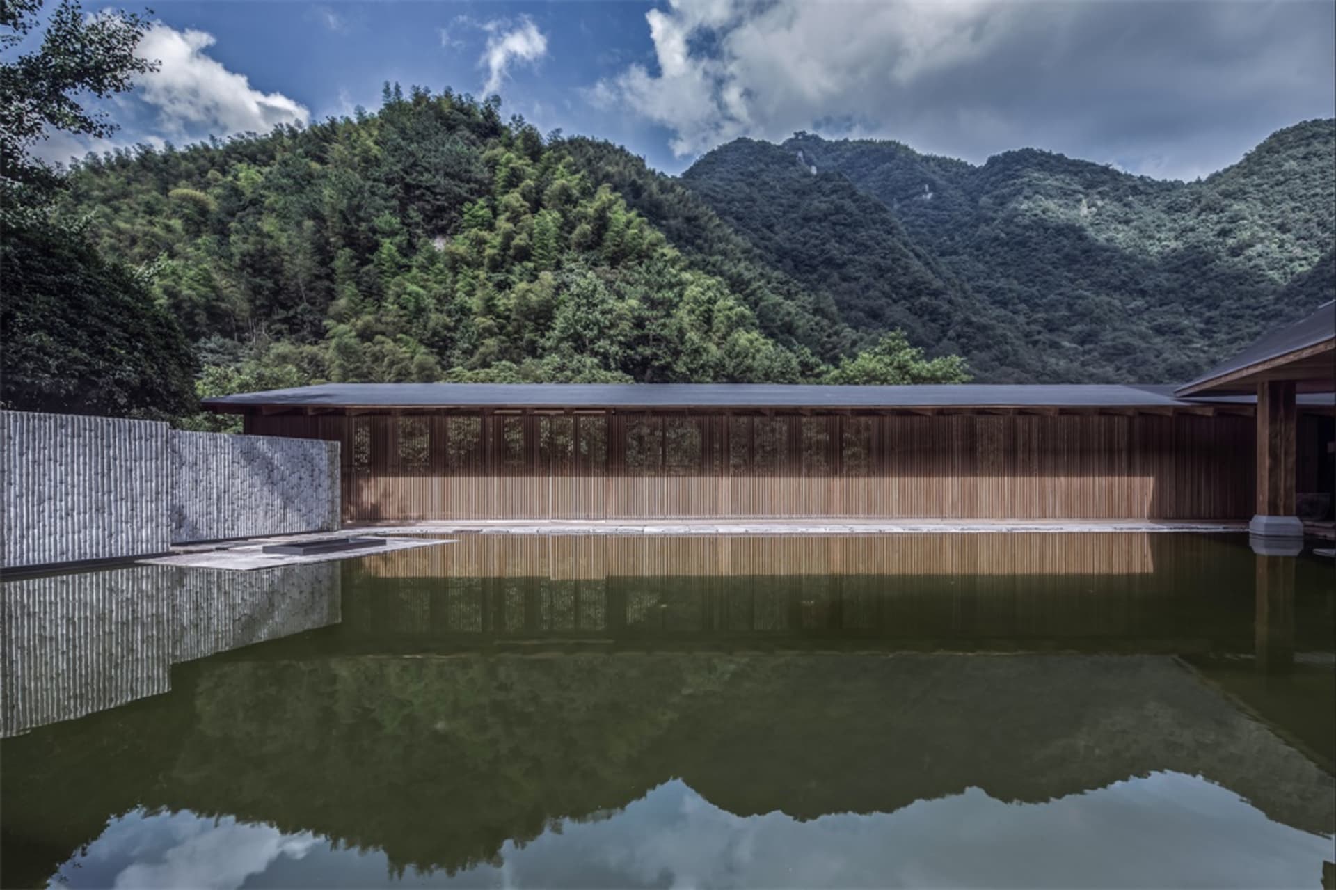 A minimalist landscape featuring a wooden building reflected in a calm pool of water, with dense green foliage and mountains