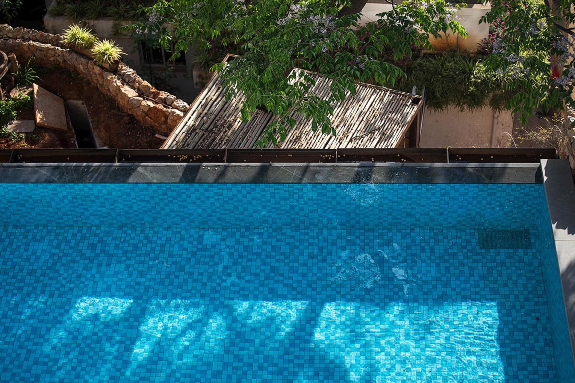 A modern poolside patio with blue tiled walls and a wooden roof structure surrounded by greenery.