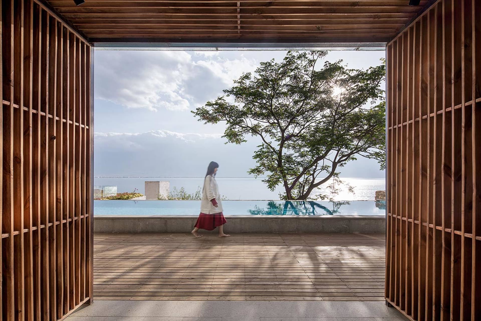 A woman walking on a wooden platform with a view of the ocean and a large tree in the background.