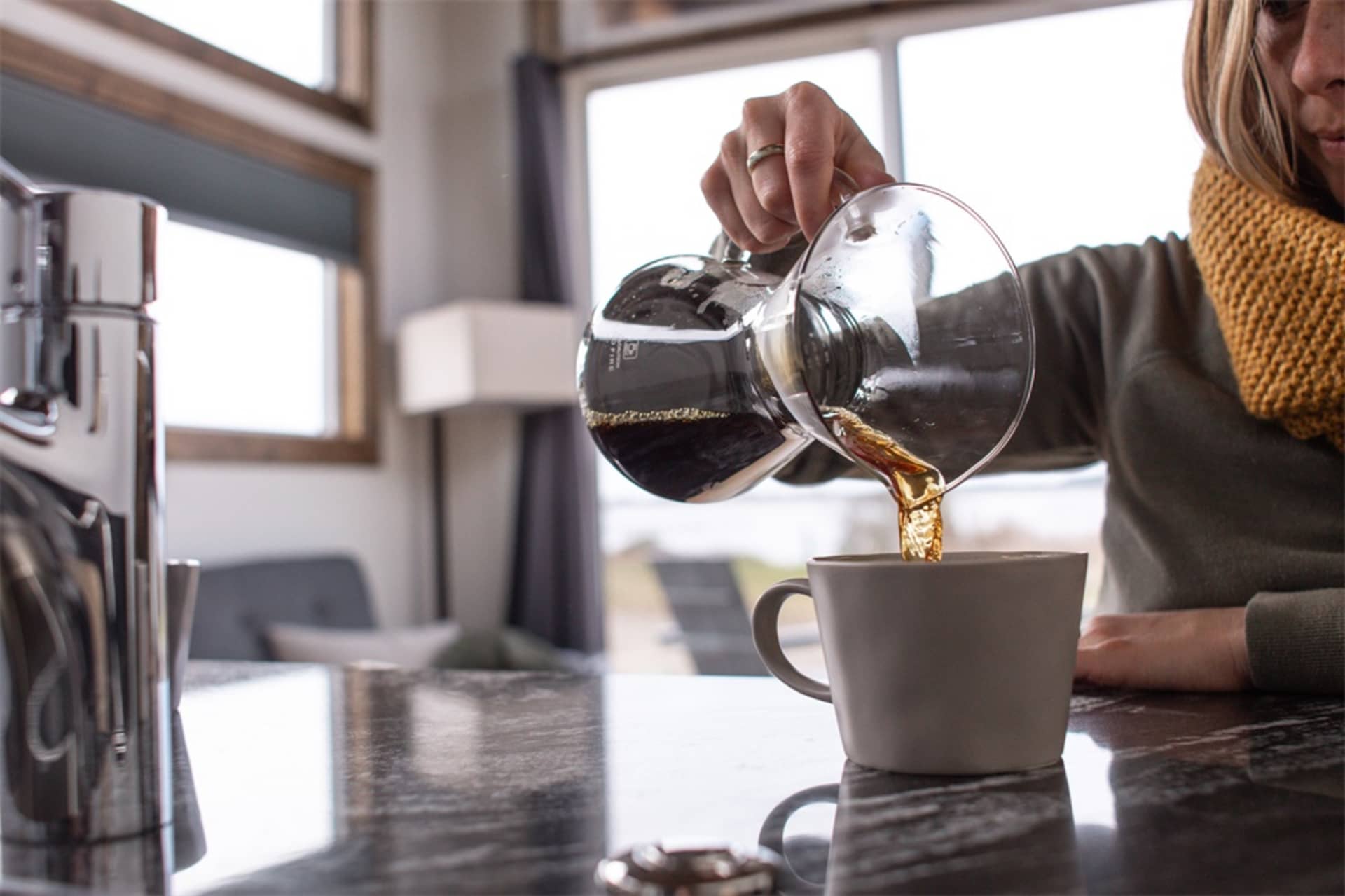 A cozy kitchen with a stone countertop and a woman pouring coffee into a mug from a glass carafe.