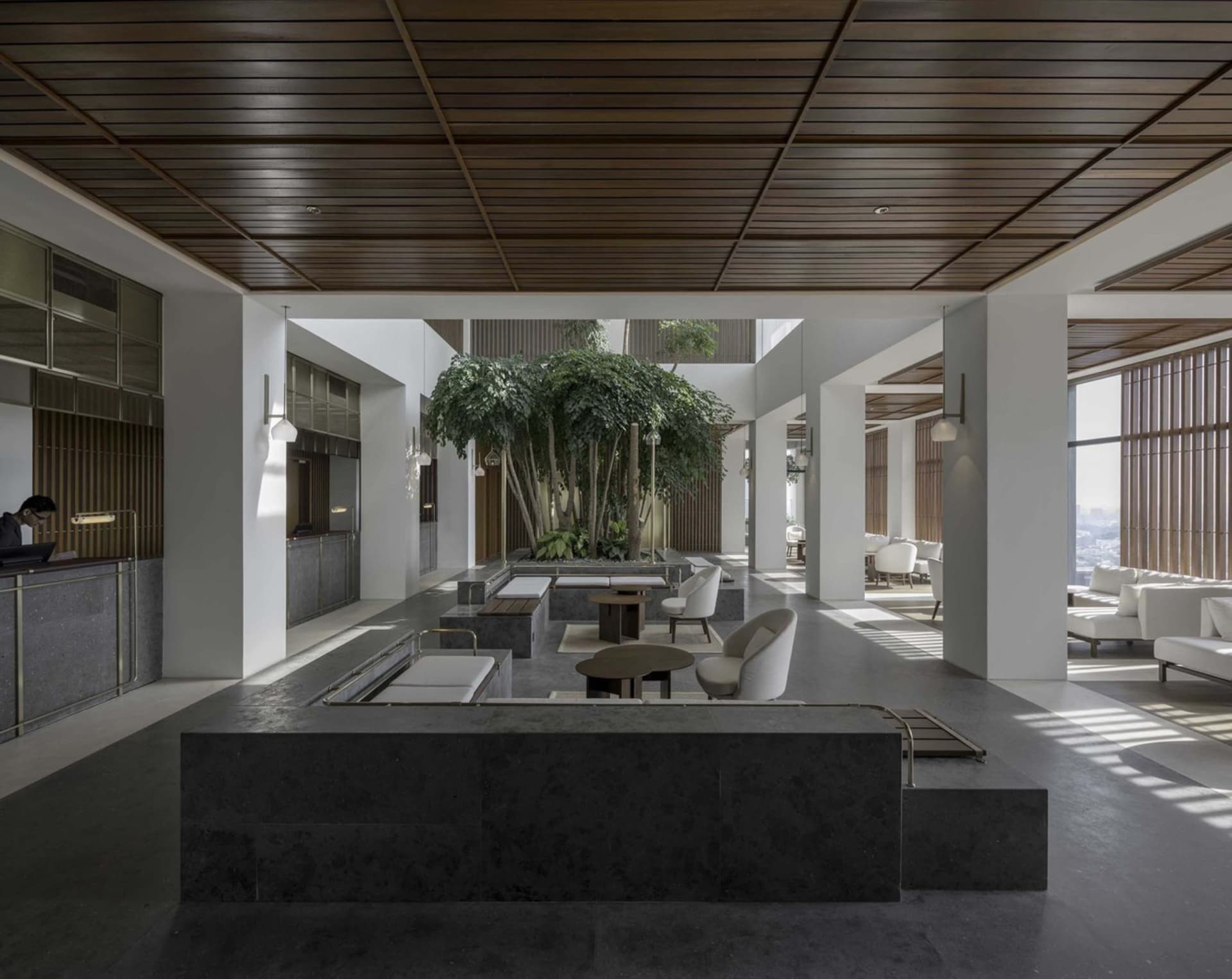 Lobby area of a modern hotel with concrete benches, white chairs, and a central greenery feature.