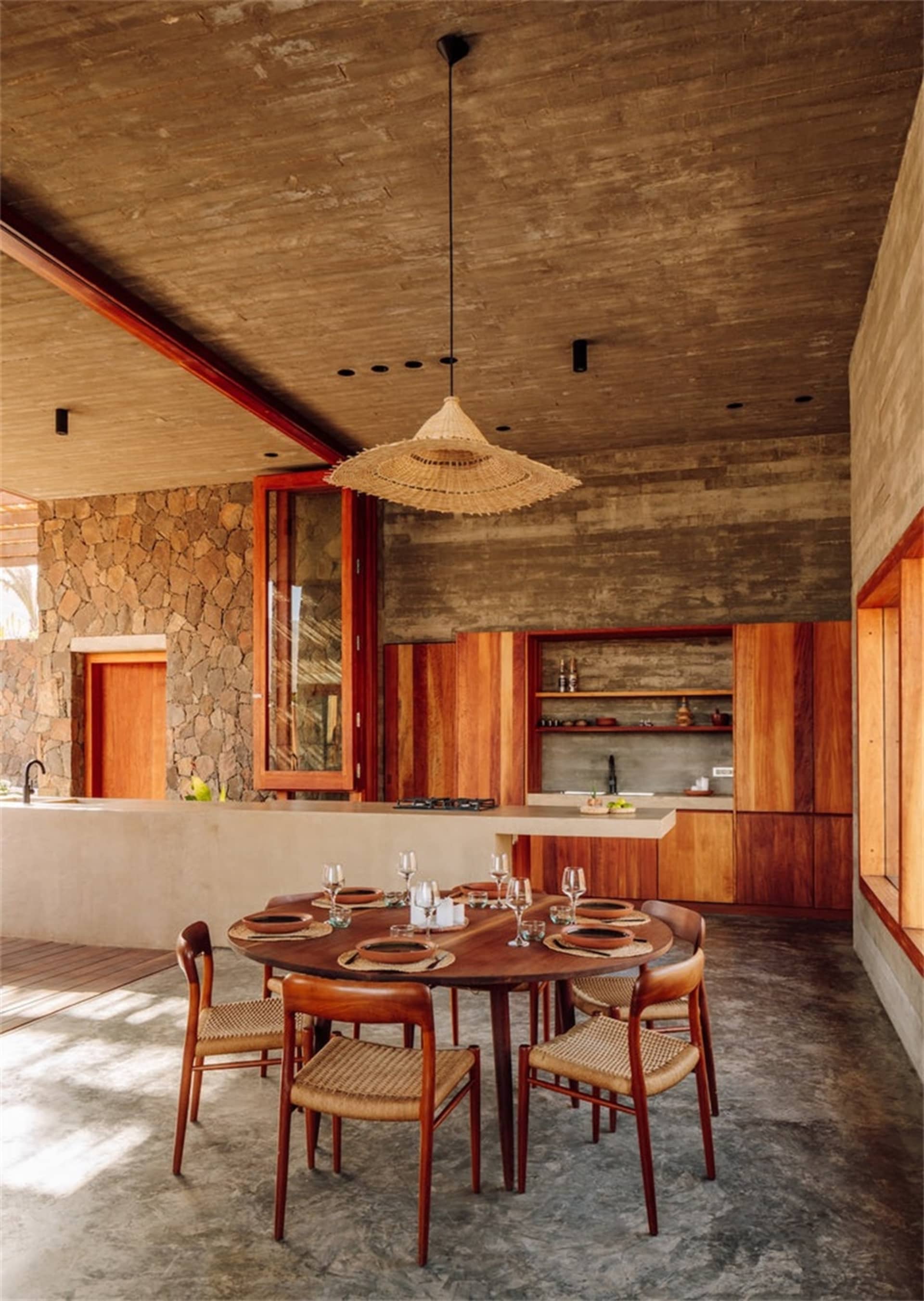 A modern dining room featuring a round wooden table, chairs with woven seats, concrete floor, stone wall, and exposed wooden