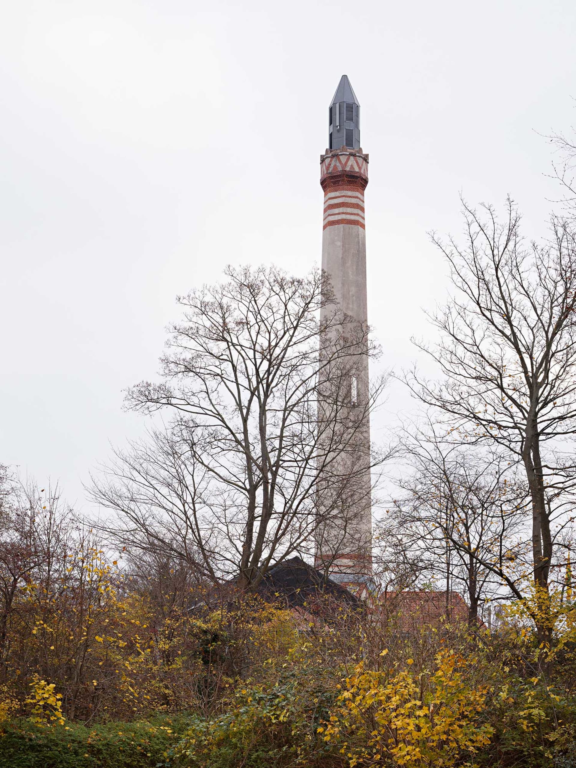 A tall, industrial concrete smokestack with red and gray stripes, surrounded by bare trees and autumn foliage.