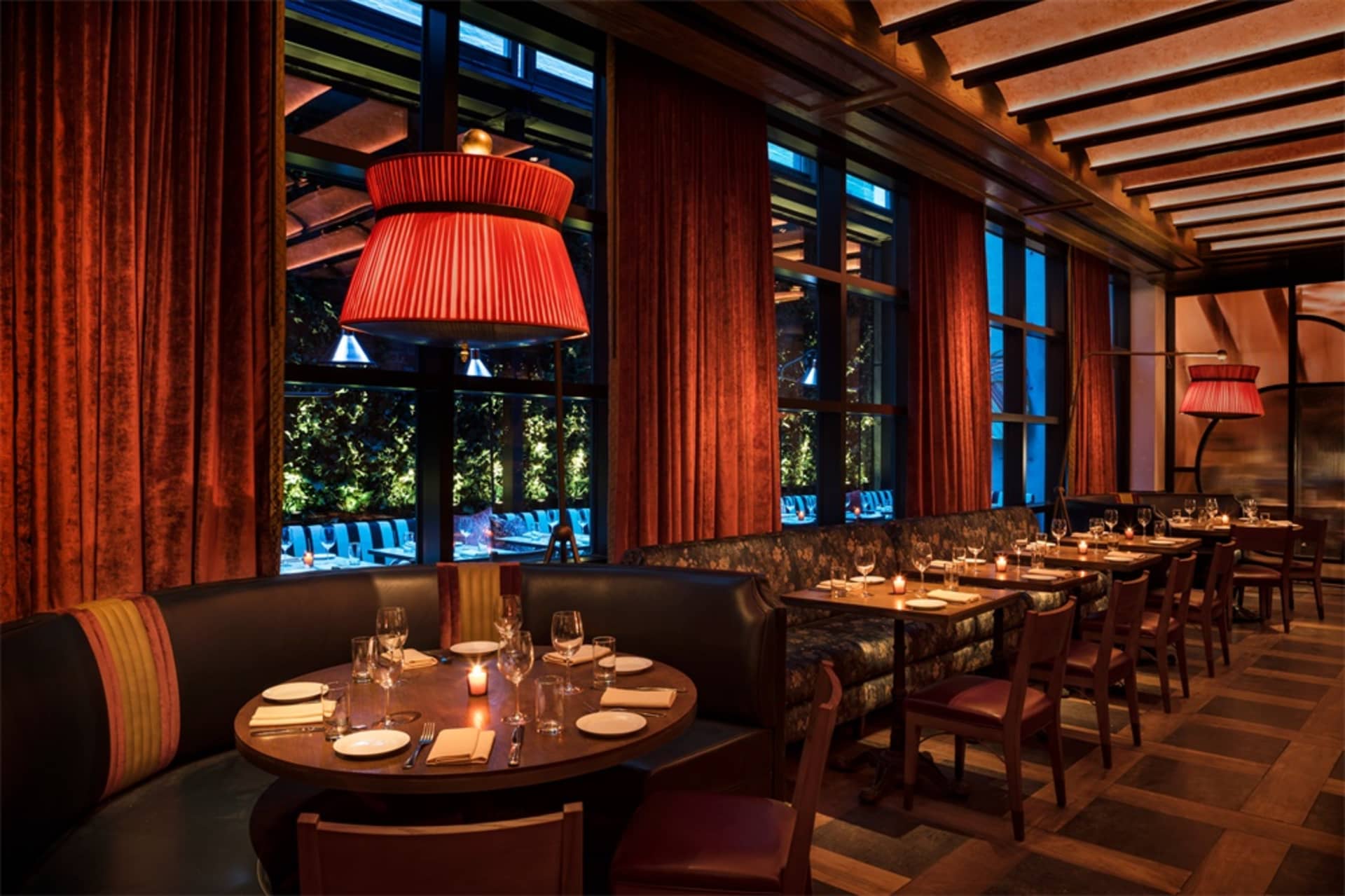 A luxurious dining room with red velvet curtains and wooden flooring, featuring round tables set for dinner with wine glasses