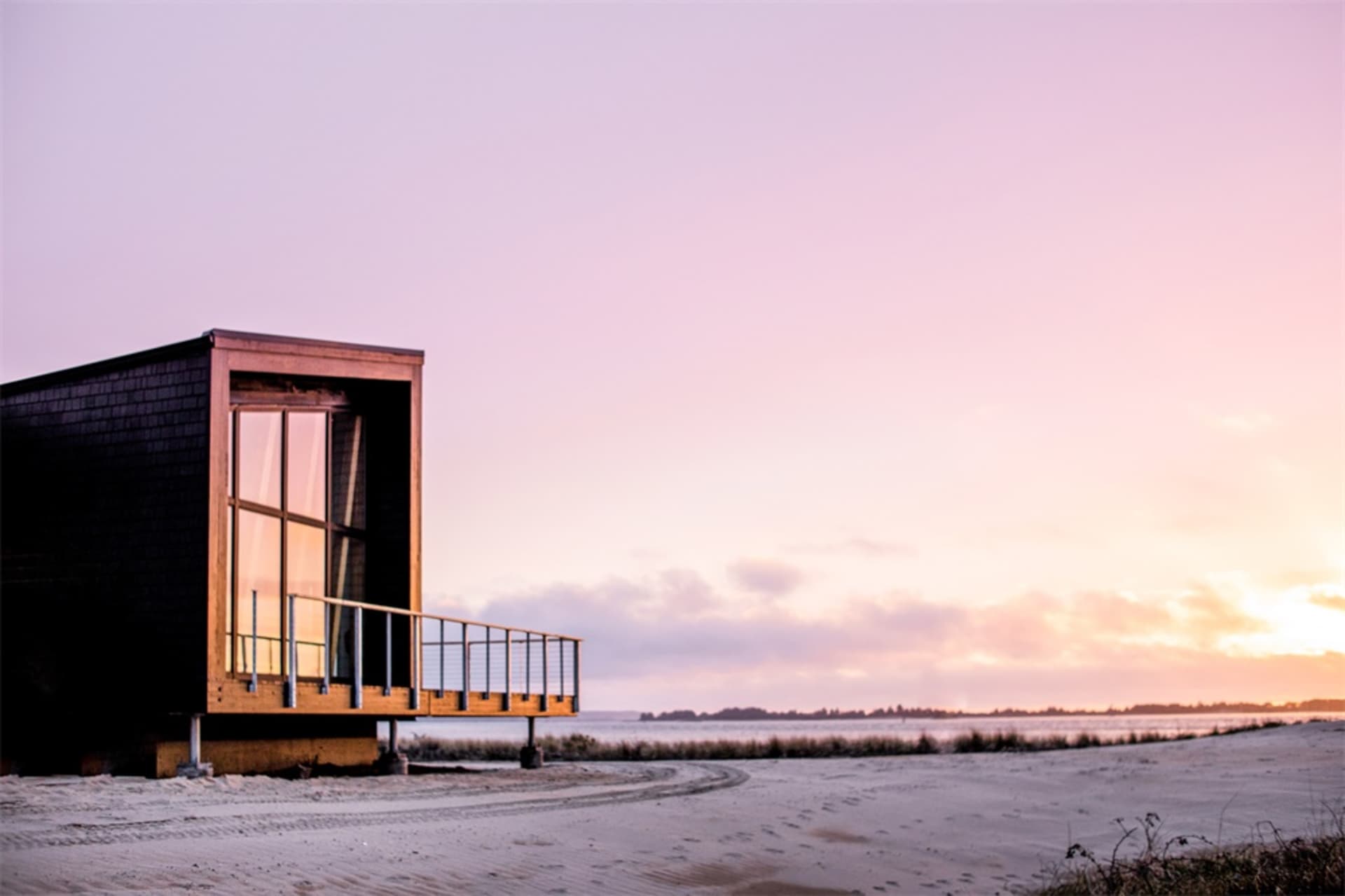 Minimalist, modern cabin with large windows and wooden exterior on a sandy beach at sunset.