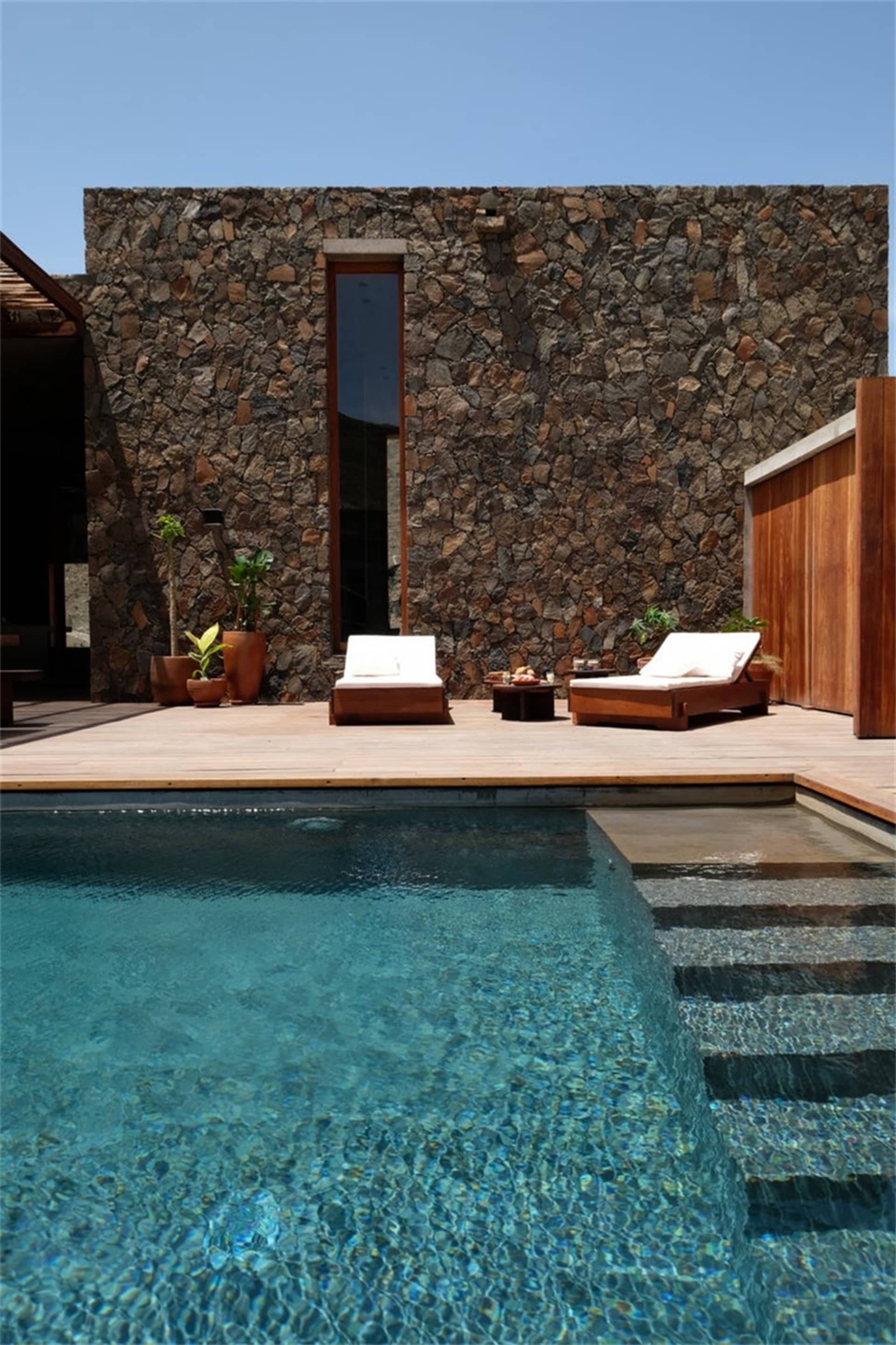 A modern pool deck featuring a stone wall, concrete pool, and lounge chairs.