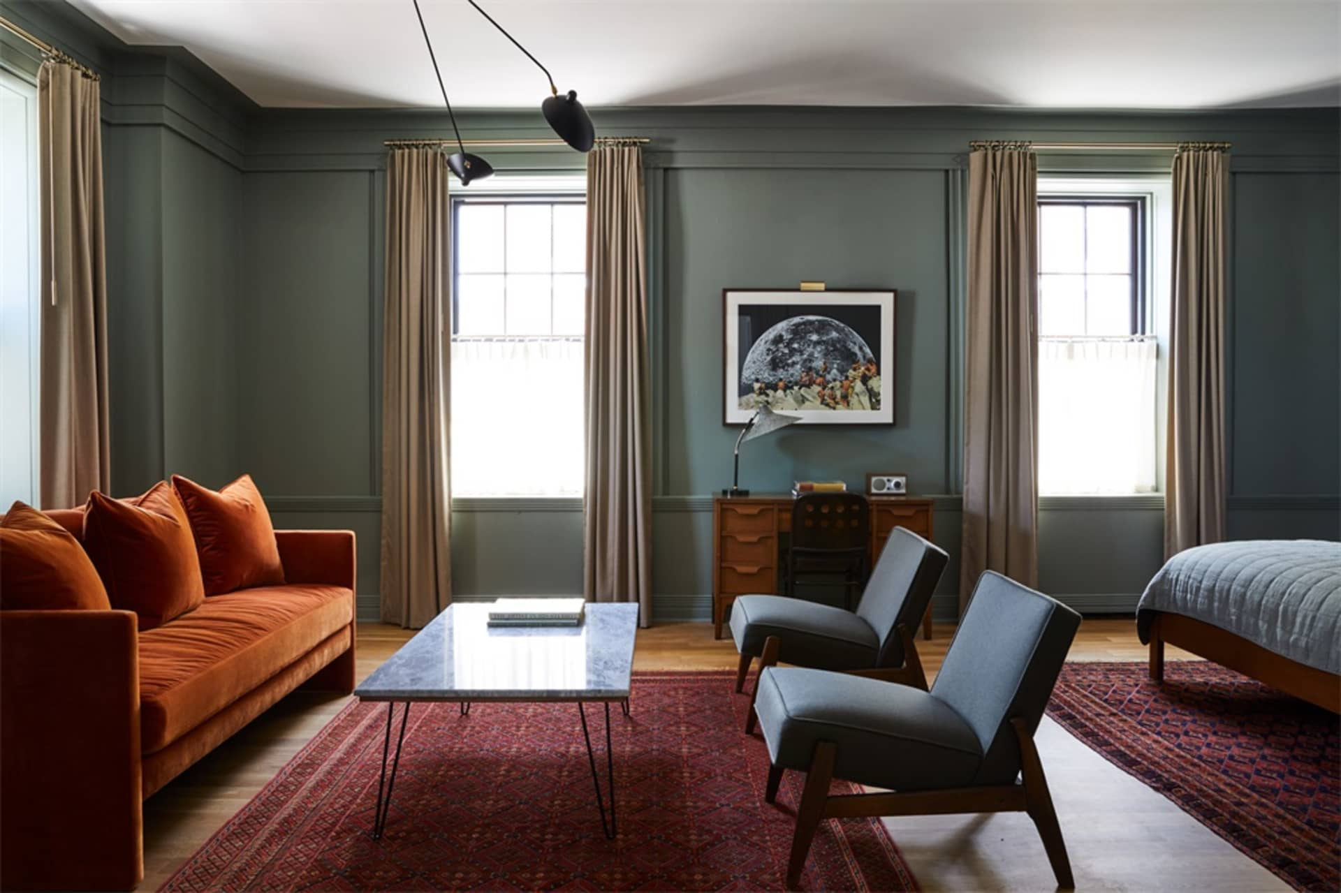 A modern living room with an orange velvet sofa, two gray armchairs, a wooden desk, and a red rug.