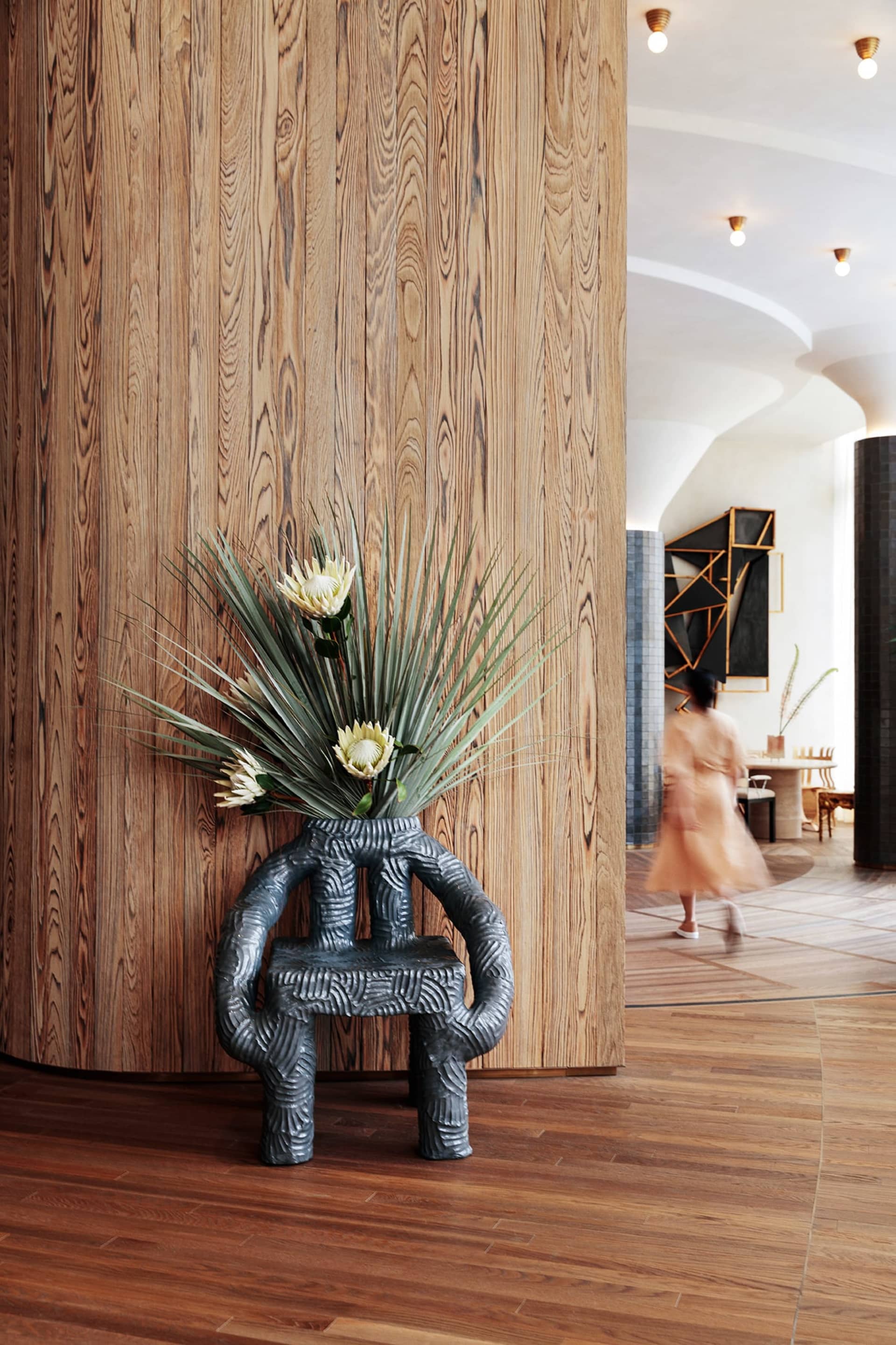 A minimalist lobby features a wooden column with a vase holding ornamental flowers and greenery, set against a textured woode
