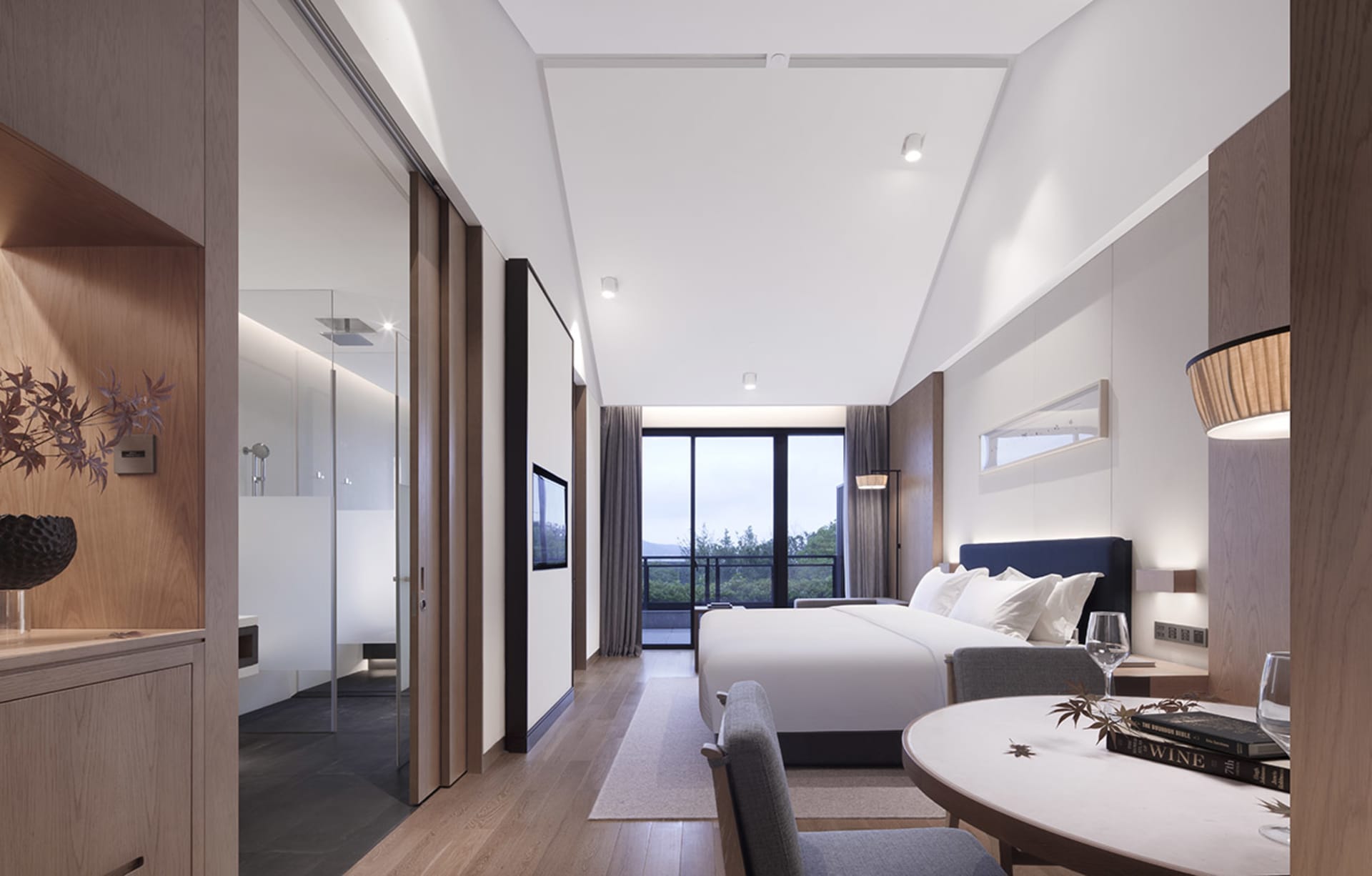 Modern hotel bedroom featuring a wooden floor, a double bed with white bedding, a round table with books and a glass, and a g