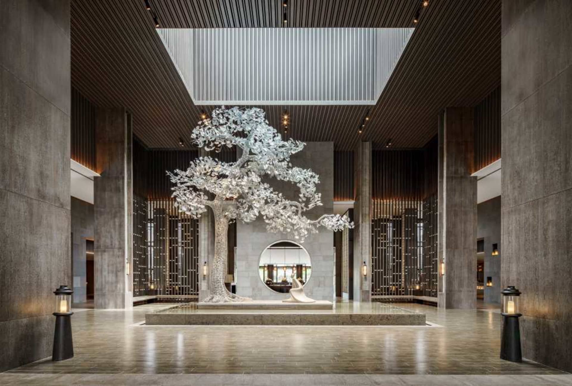 現代酒店大廳內,一座大型金屬樹雕塑置於石質基座上,周圍有高聳柱列與天窗。