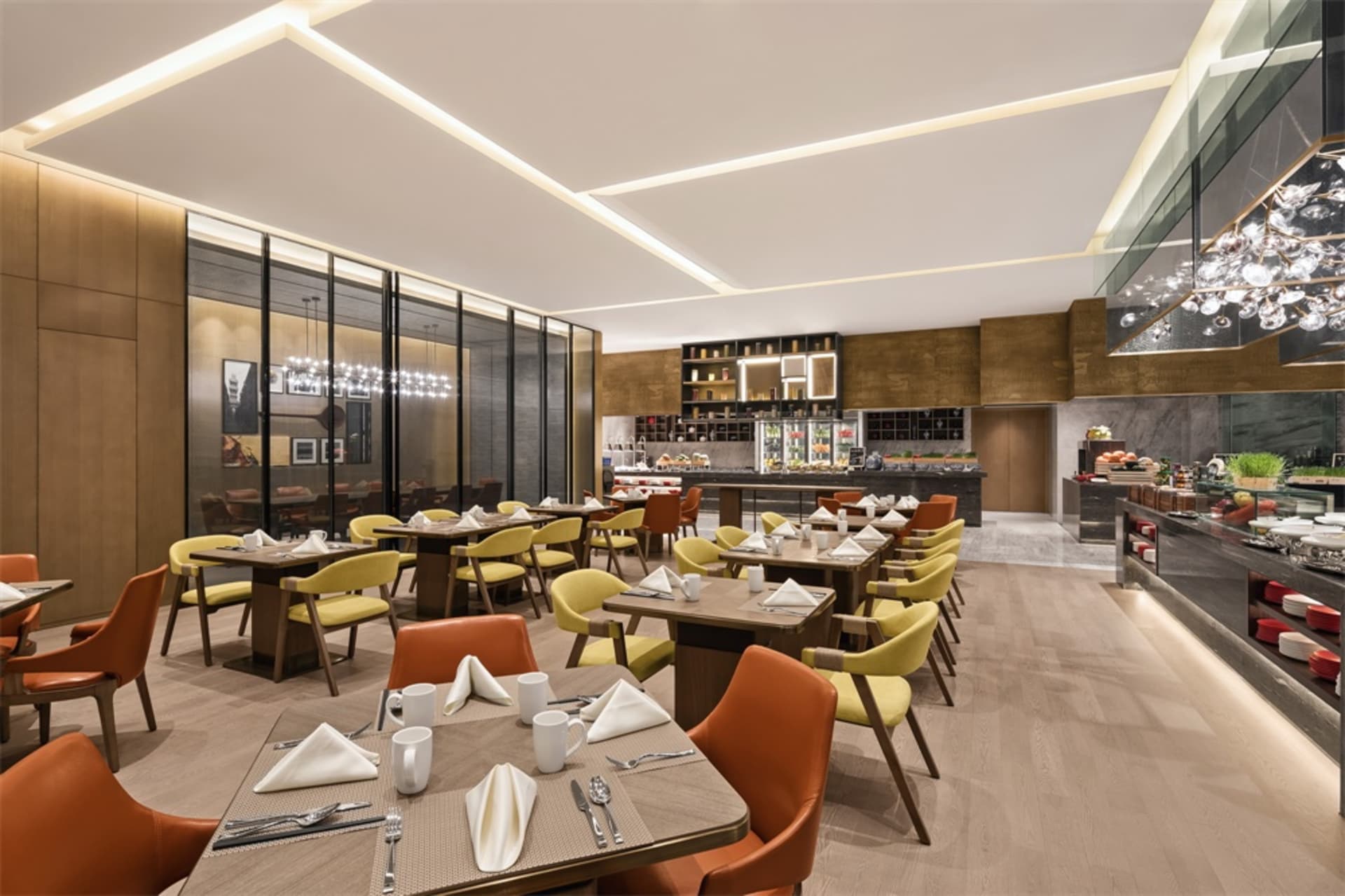 A spacious, modern restaurant interior with wooden tables, yellow chairs, and a well-lit kitchen area.
