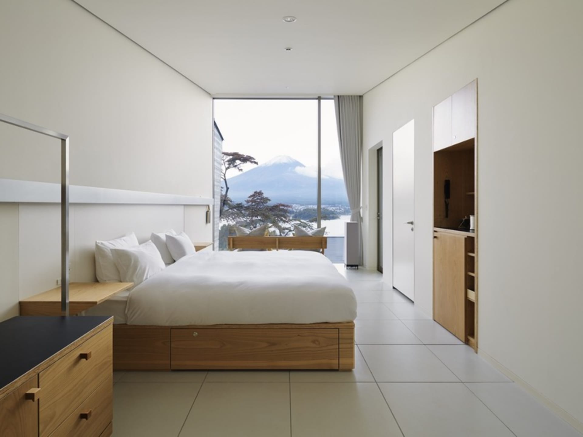 A modern minimalist bedroom features a large bed with white bedding, a glass window, and wooden furniture with a view of a mo