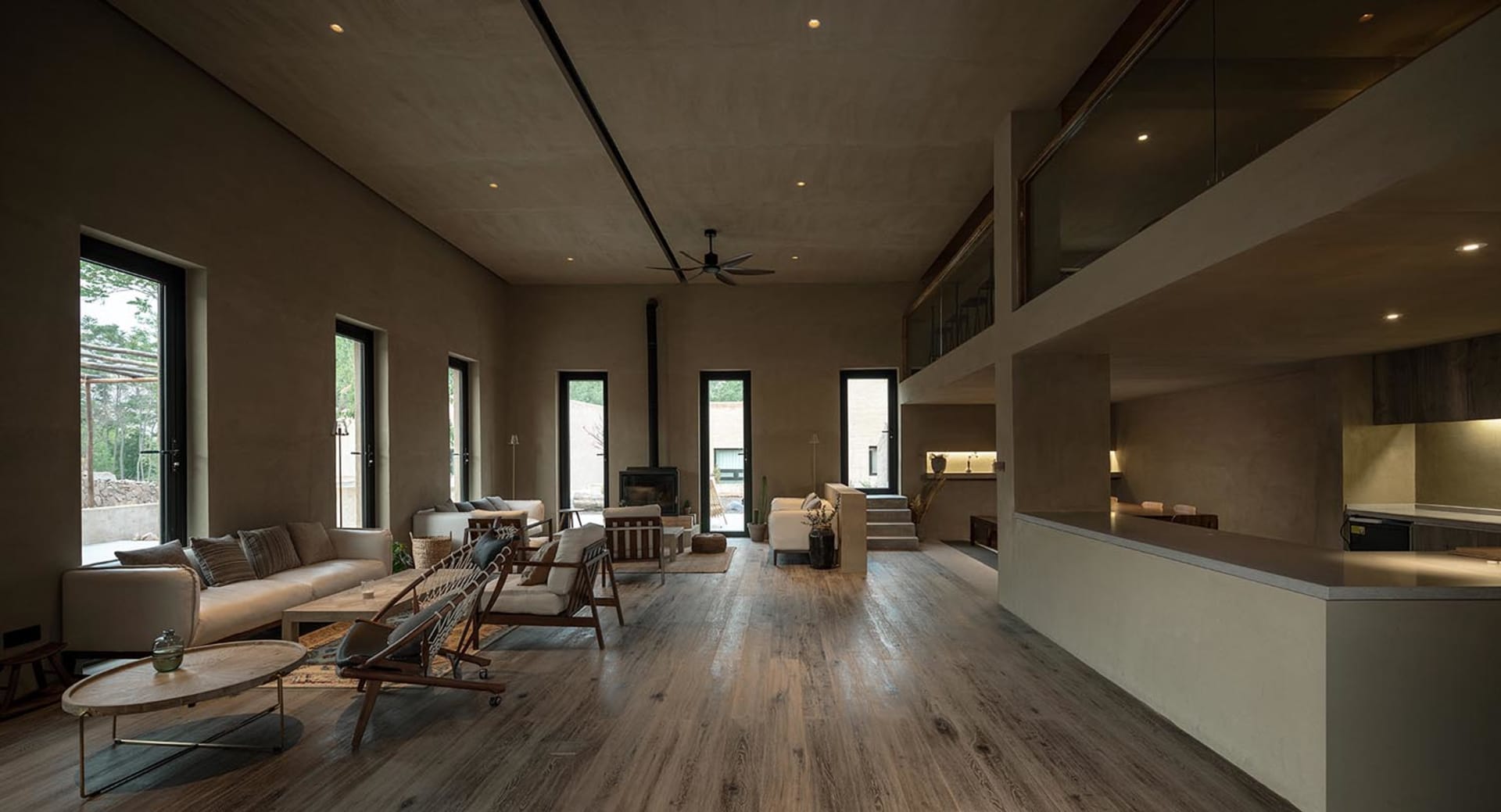 A modern living room with concrete flooring, wooden furniture, and a central fireplace. The space is illuminated by natural l