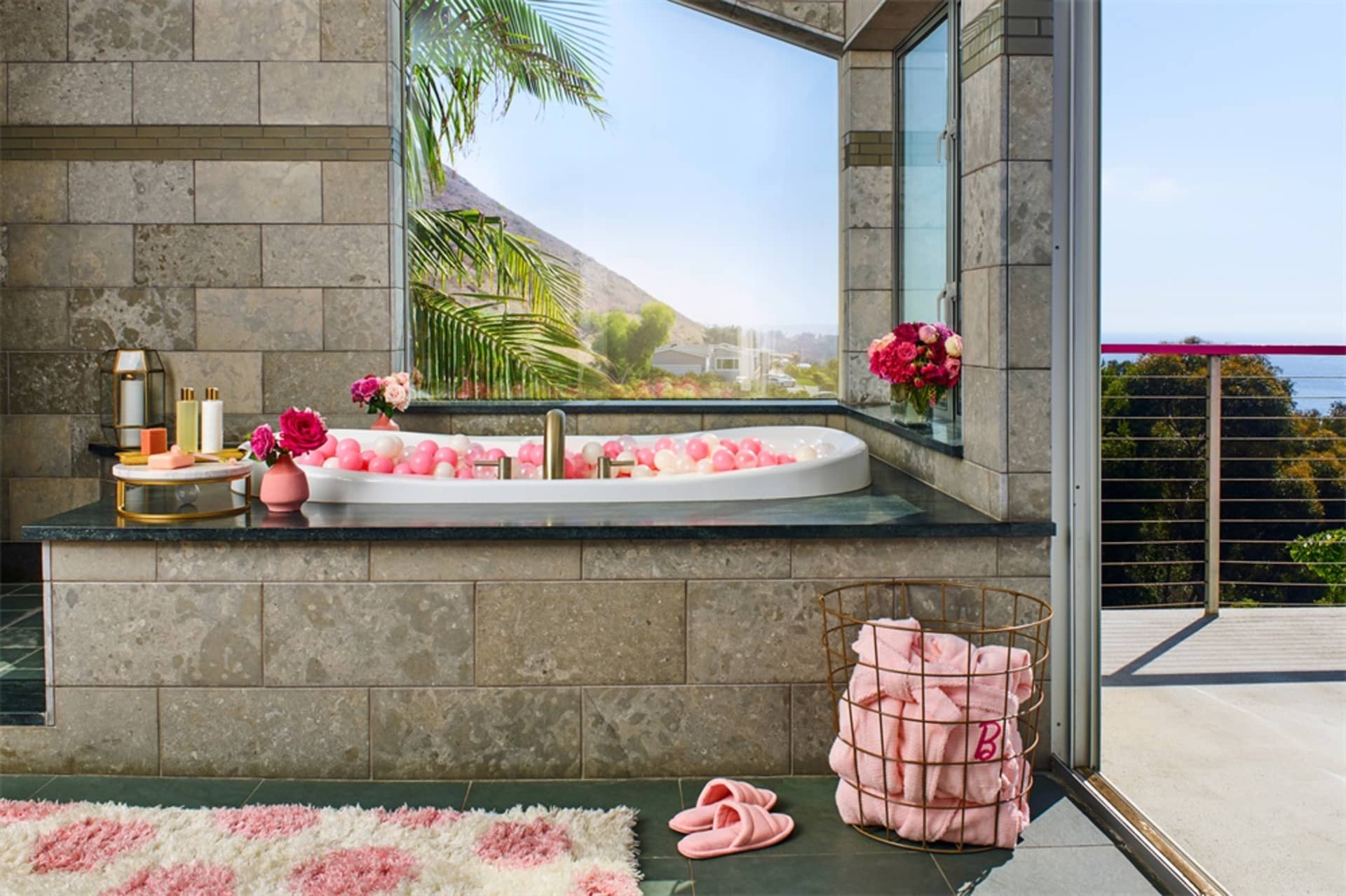 A modern spa bathroom with stone tile walls, a large window overlooking a mountain view, a bathtub filled with colorful balls