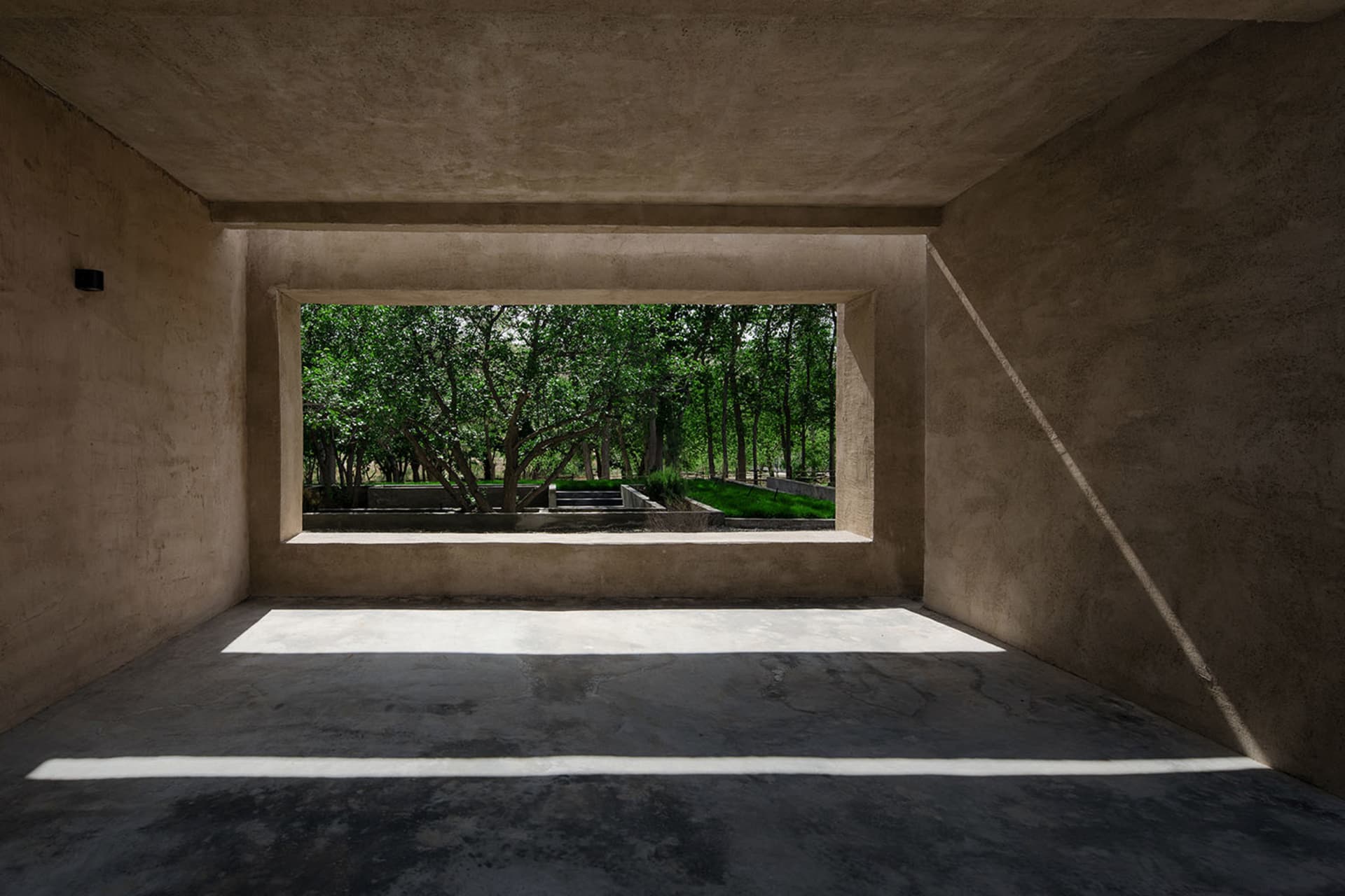 Modern interior space with concrete walls and floor, framed by a large window showcasing a green outdoor area with trees.