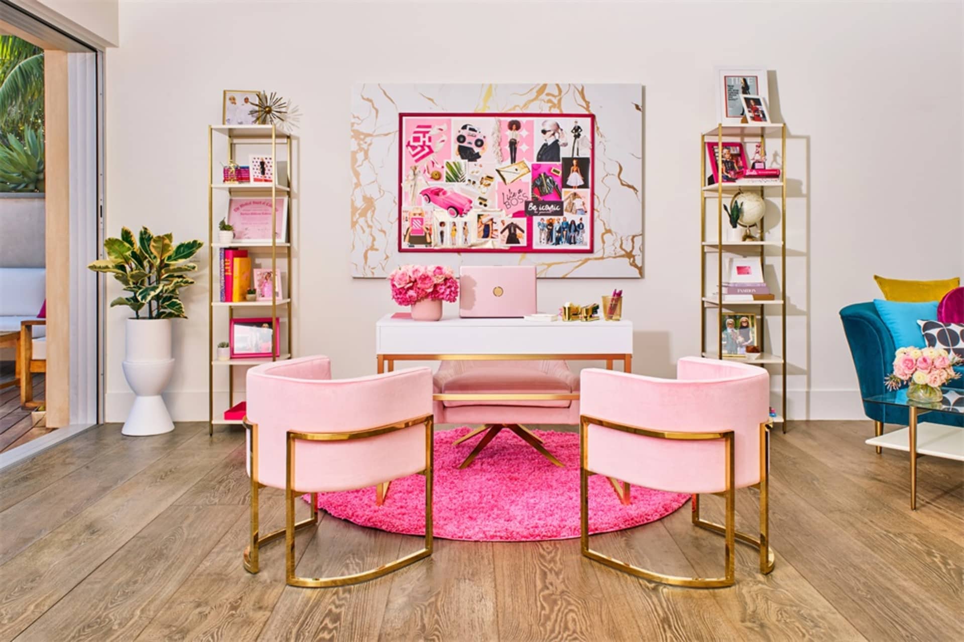 Modern office with pink chairs and golden accents, featuring a white desk and metallic shelving units with decorative items.
