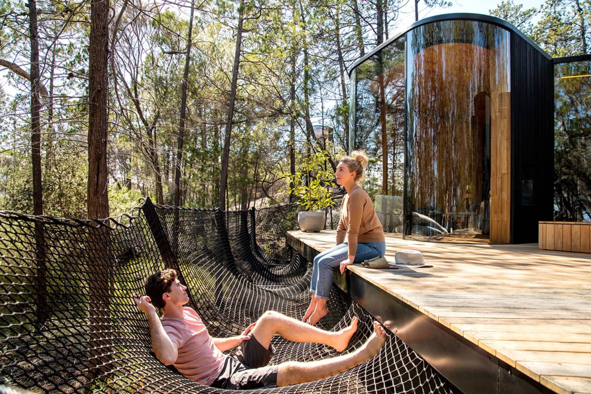 A man relaxing in a mesh net chair and a woman sitting on a wooden deck with modern architecture in the background.