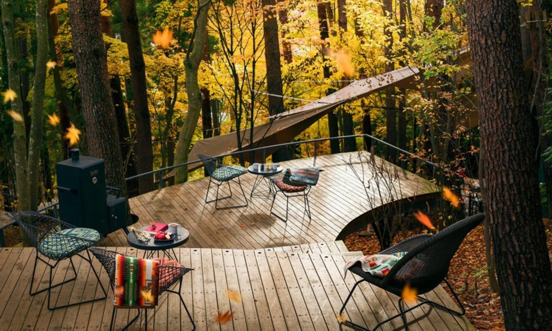 A deck surrounded by autumn foliage with wooden furniture and a grill.