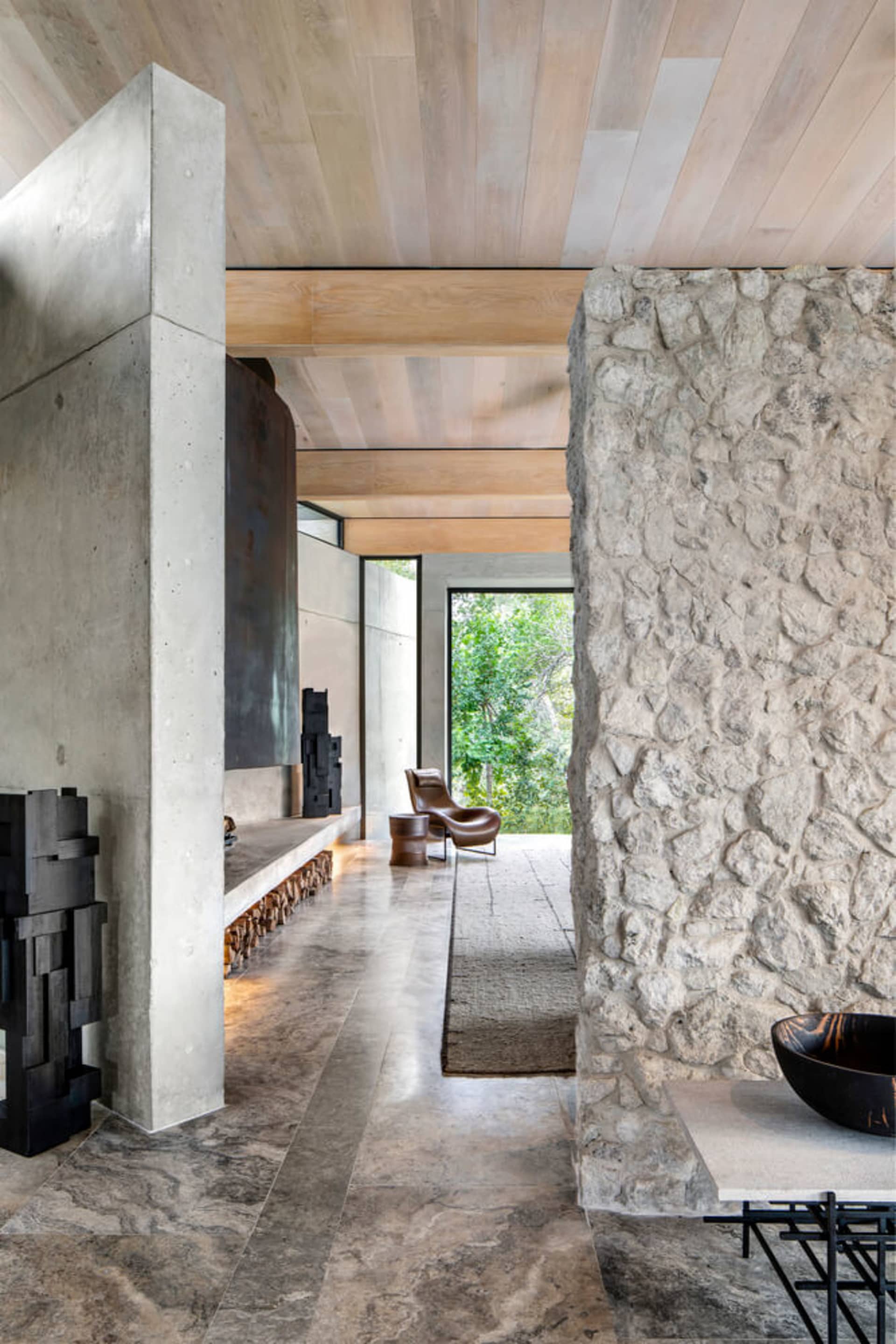 A modern living room featuring concrete walls, floor, and ceiling, with a cozy fireplace and a view of greenery outside.