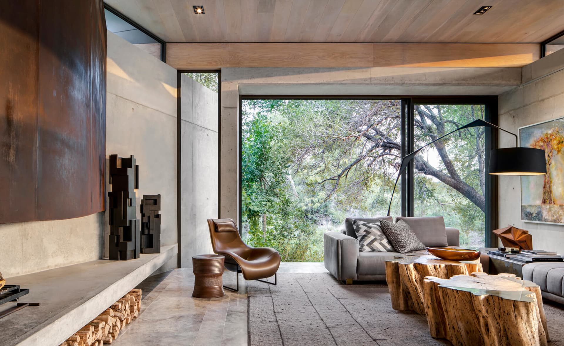 Modern living room featuring a concrete fireplace, wooden floor, metal sculpture, and glass coffee table.